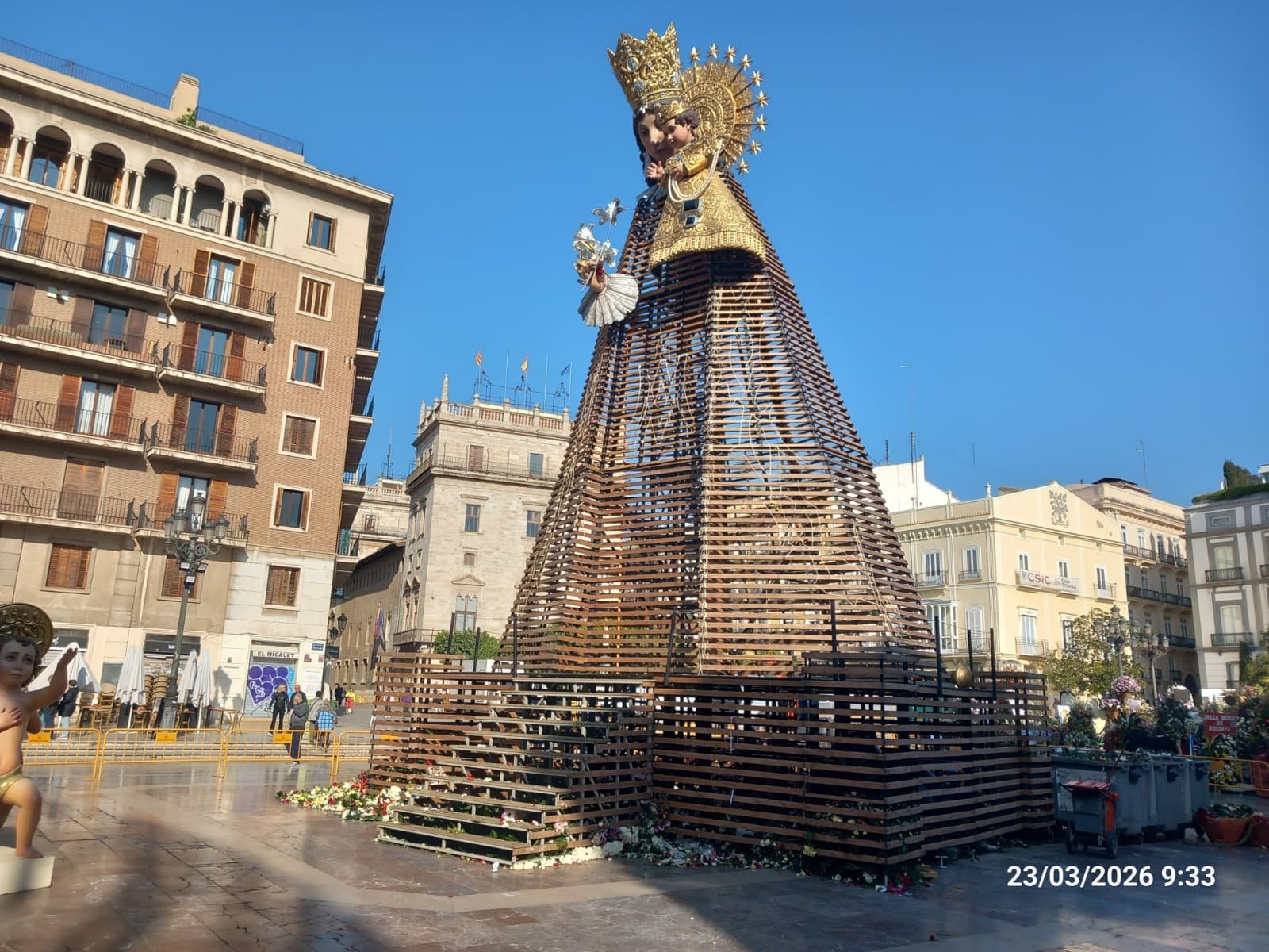 Estructura de madera en forma de Virgen coronada con el Niño en una plaza, flores en la base y edificios alrededor.