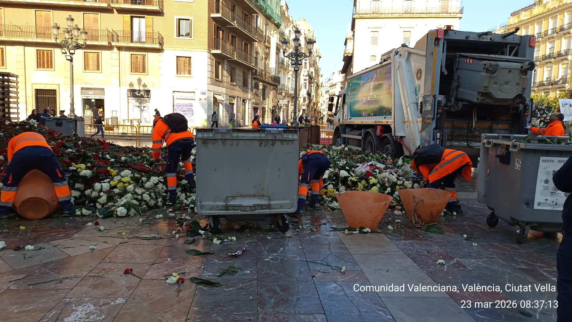 Operarios con chalecos naranjas recogen flores y restos en una plaza junto a un camión de recogida y varios contenedores.