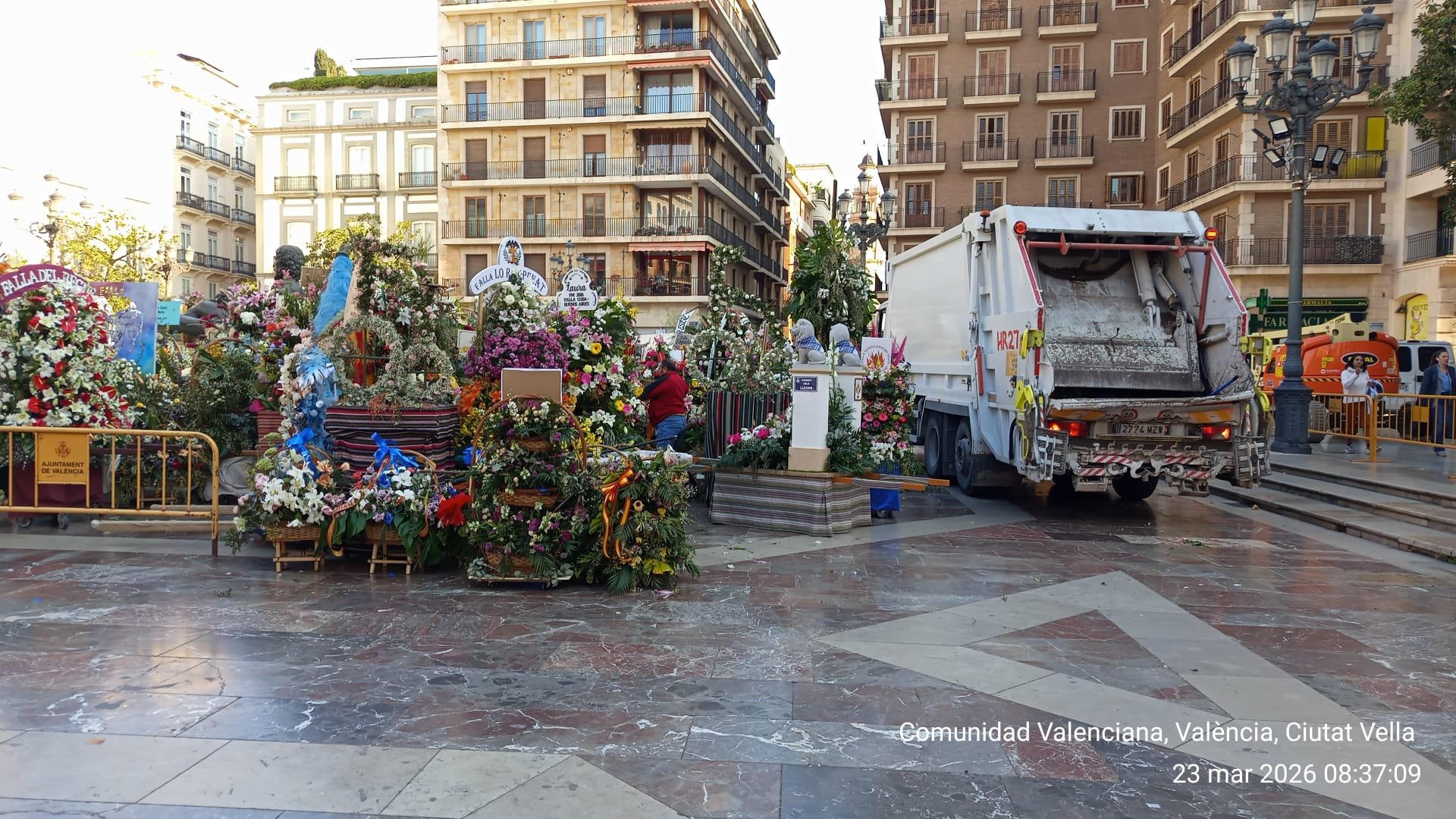 Ofrendas florales coloridas y figuras en una plaza de Valencia, con un camión de basura aparcado junto a ellas.