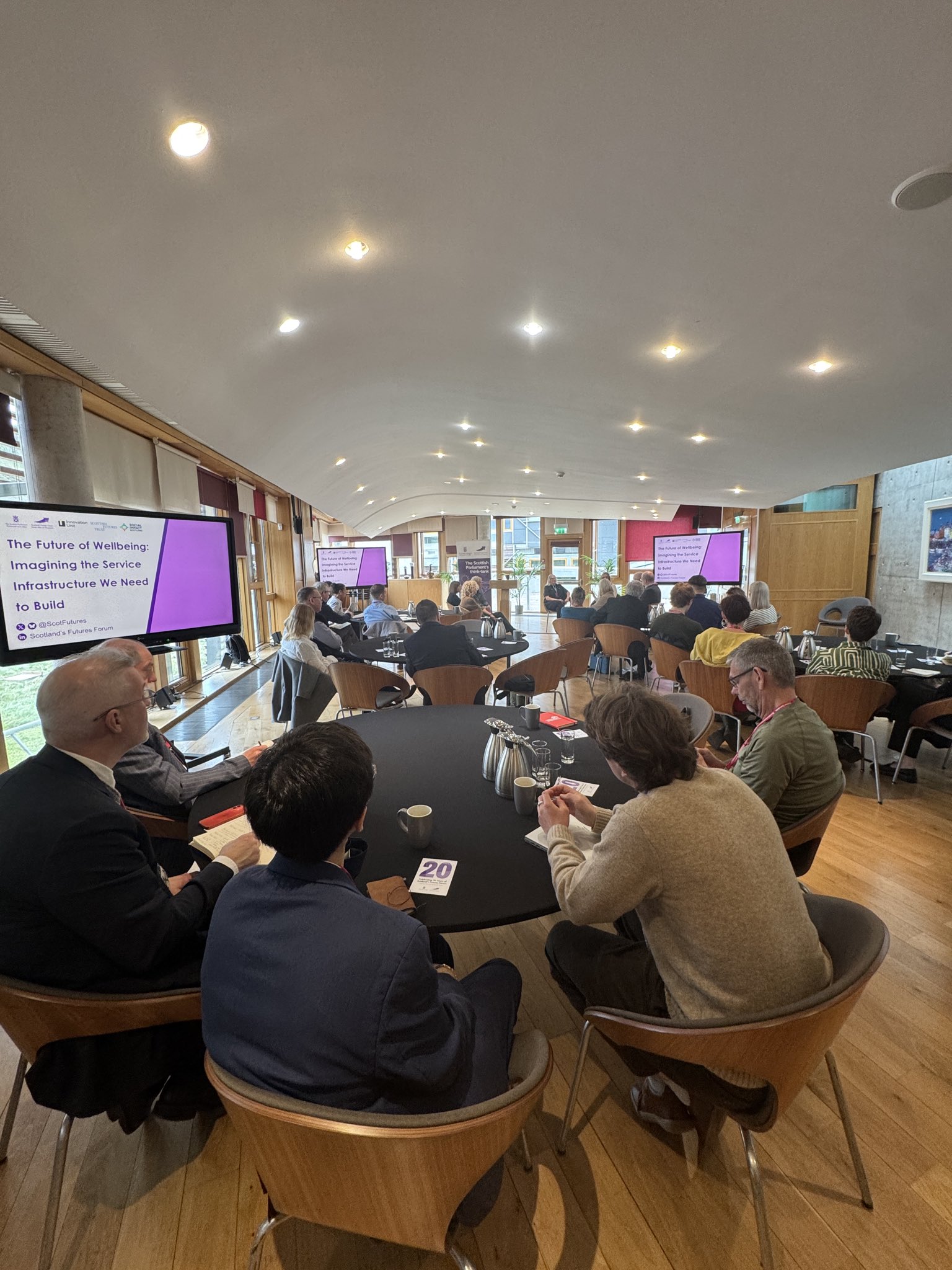 Room filled with people sitting around round tables, screens showing slide saying “The Future of Wellbeing: Imagining the Service Infrastructure We Need to Build”
