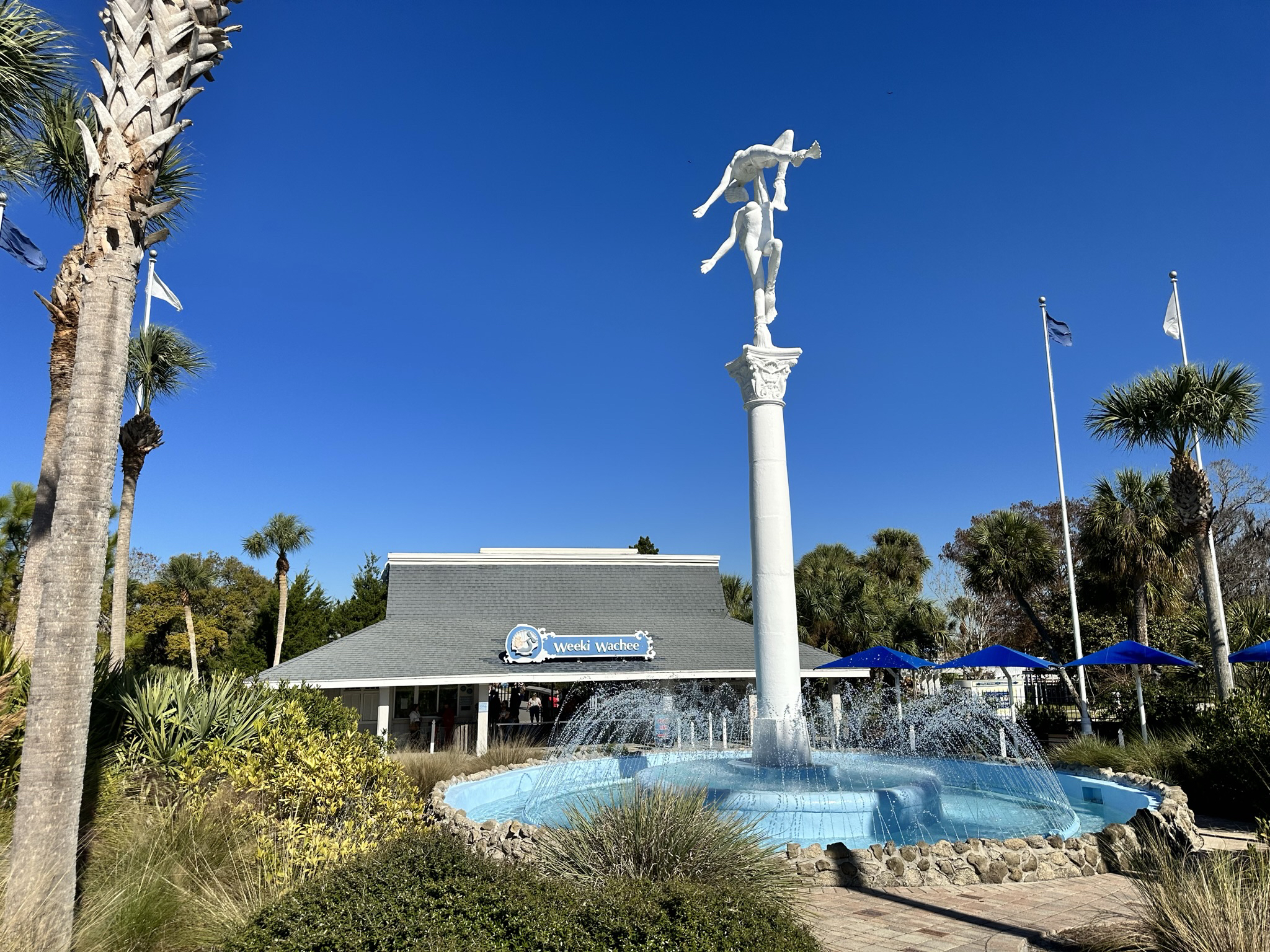 The image features the entrance of Weeki Wachee Springs State Park in Hernando County, Florida, showcasing a tall fountain with a sculpture of two people. Palm trees and greenery surround the area, and the sky is clear and blue.