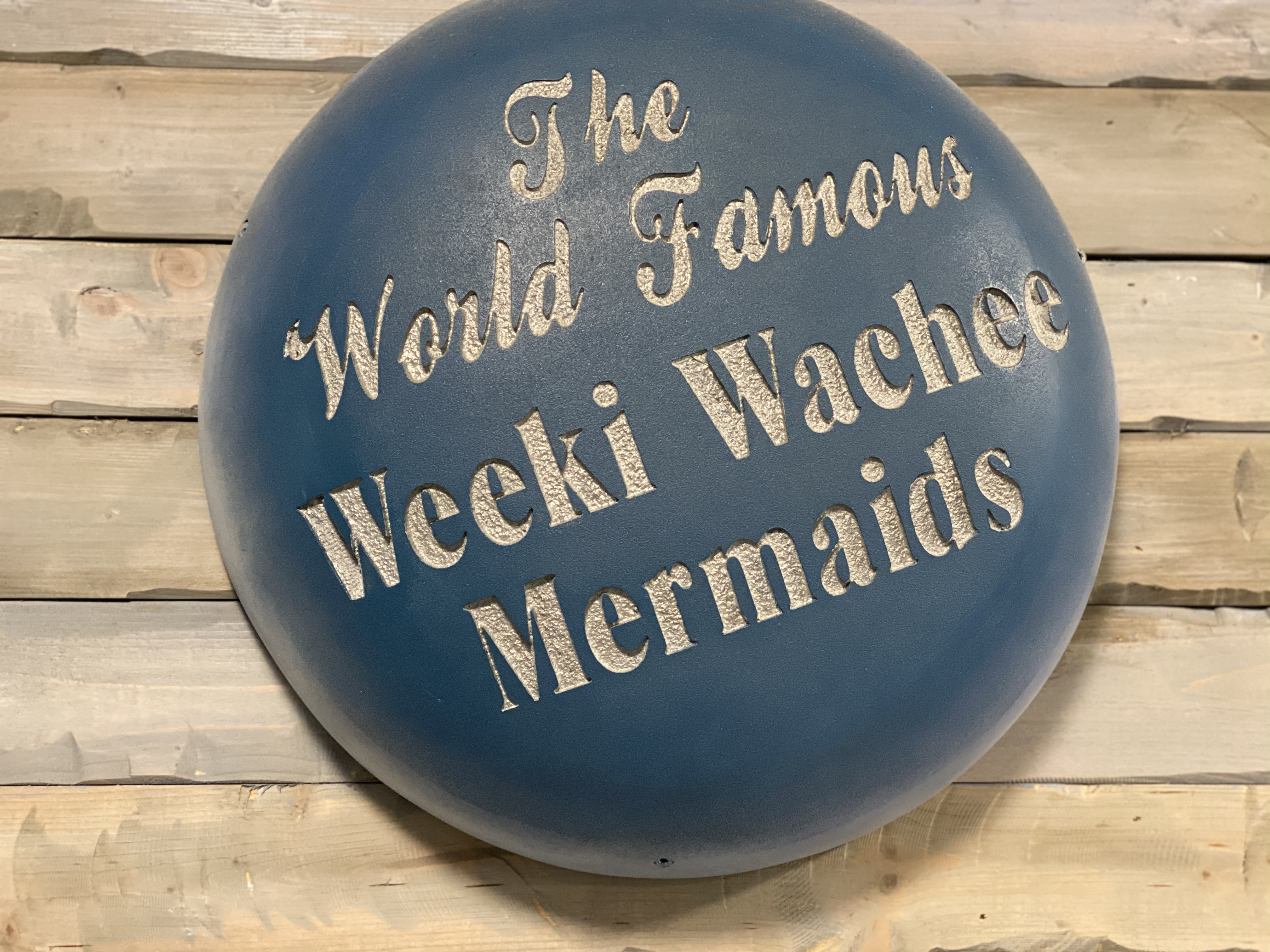 A blue oval sign with white text reads: "The World Famous Weeki Wachee Mermaids." The background is wooden planks.