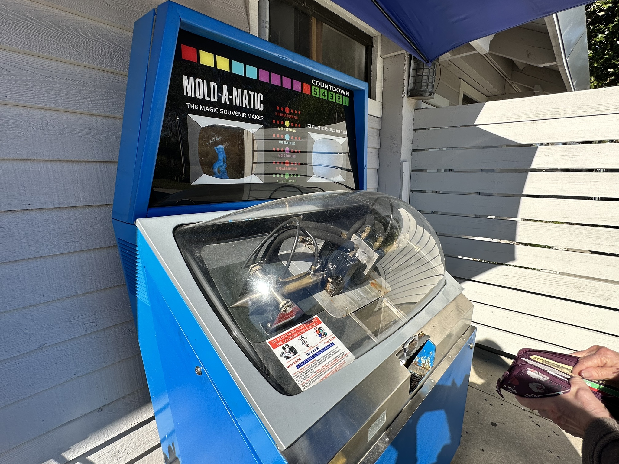 A blue Mold-A-Matic machine is outdoors against a wooden wall. It has a clear dome displaying mechanical components inside that create mermaid wax figures.