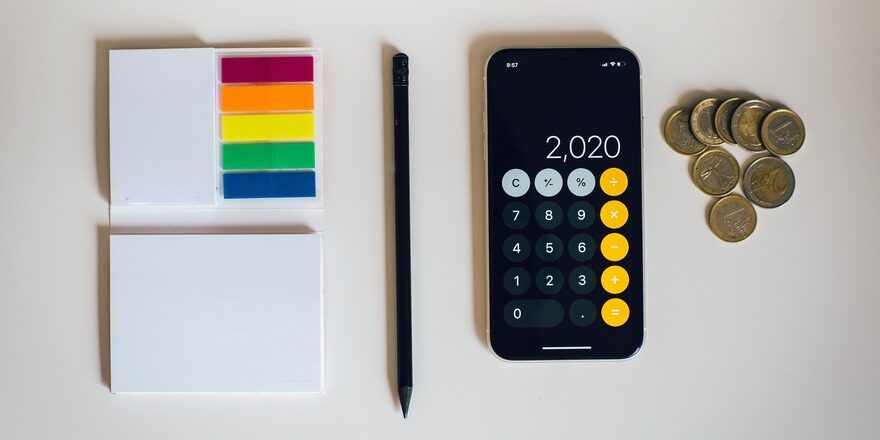 A white pad of paper, colored page flags, a black pencil, and a smartphone displaying the calculator app sit next to coins.