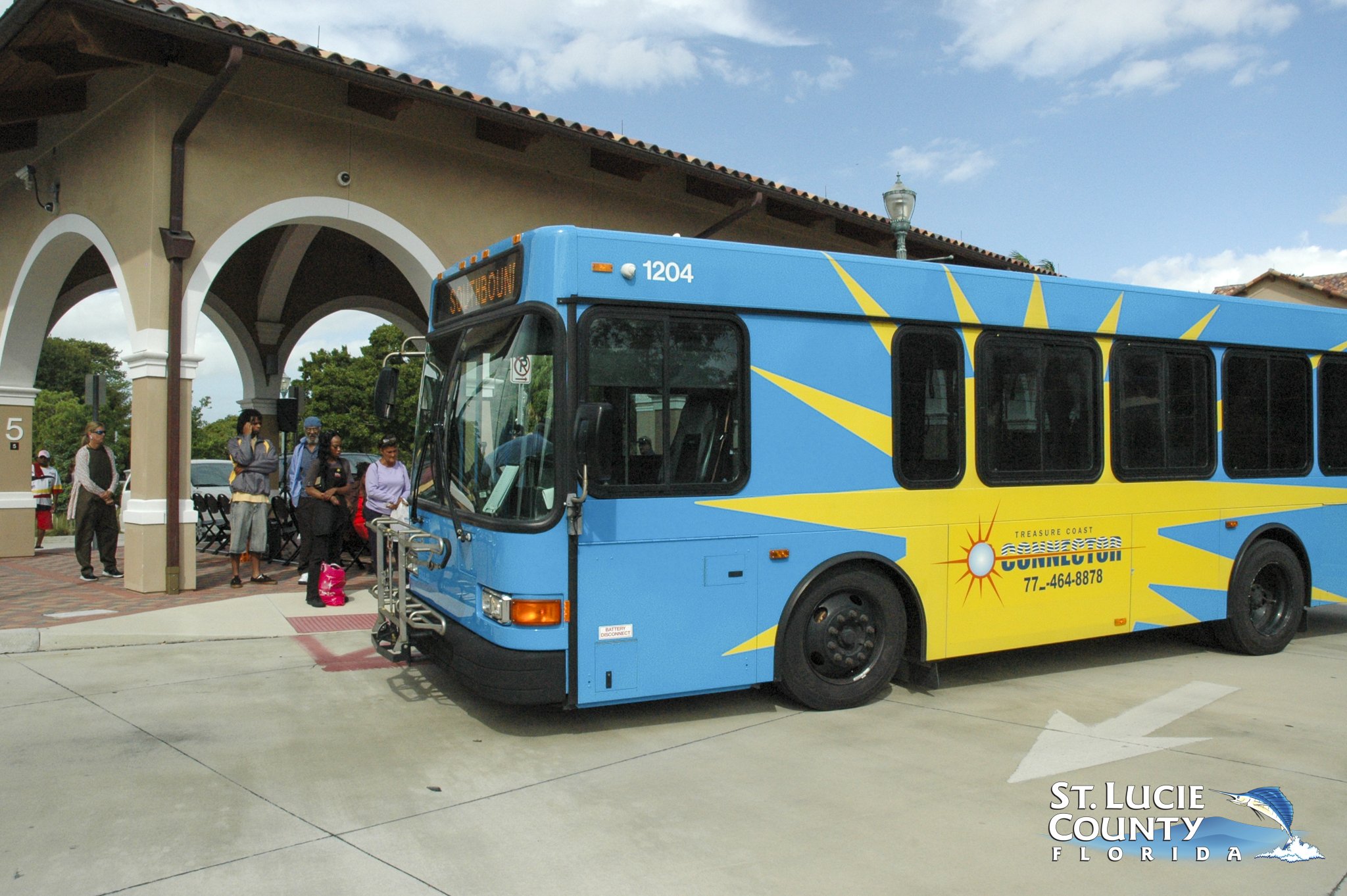 Bright blue public bus with yellow sunburst design parked at a station where passengers wait under an arched shelter.