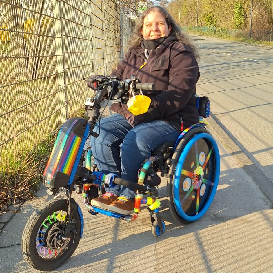 Ich sitze in meinem Rollstuhl und habe mein Zuggerät dran.
Ich werde voll von der Sonne angestrahlt und mein buntes Gespann leuchtet ebenfalls in der Sonne.
Ich lächle glücklich.