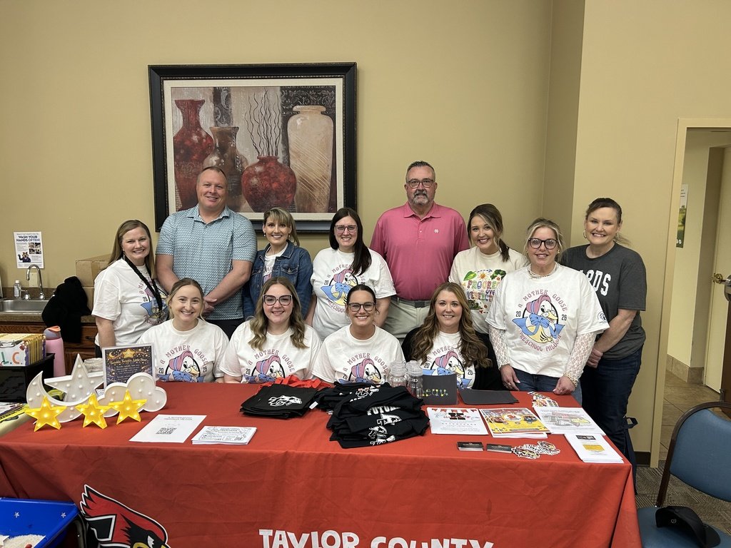 Group photo indoors: twelve adults stand and sit behind a table with a red Taylor County tablecloth, flyers, shirts, and decorations. A framed vase artwork hangs on the wall behind them.