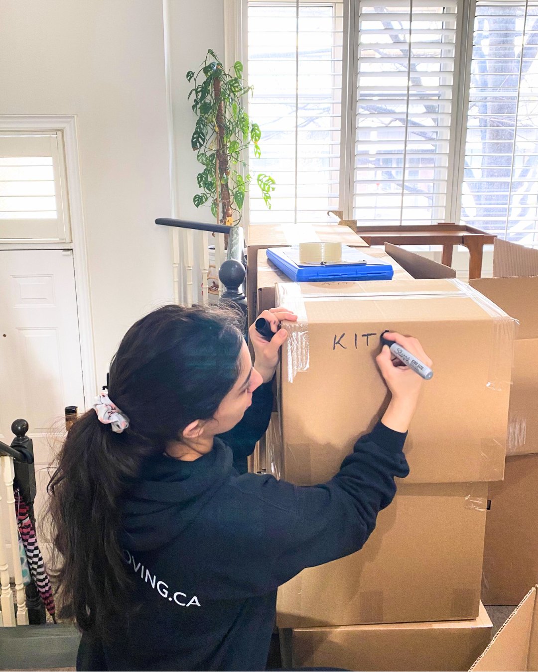 A Six Moving packer writing "KITCHEN" on a cardboard box with a marker. The boxes are stacked in a well-lit room with white walls and large windows.