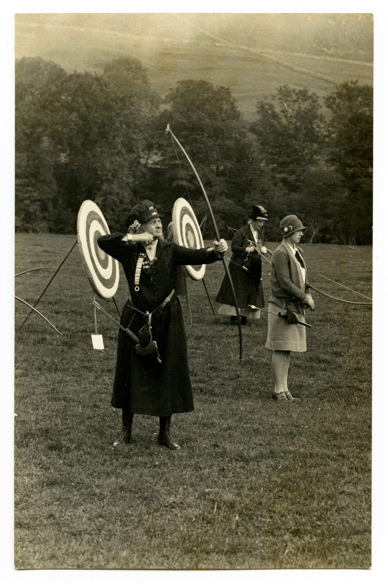 Black and white photograph of women practicing archery outdoors, with one woman drawing a bow in front of circular targets and others standing nearby on a grassy field with trees in the background.