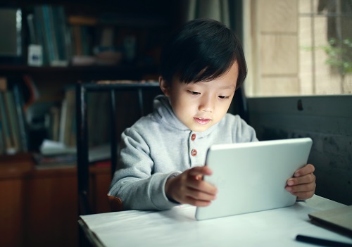 Child wearing a gray hoodie intently using a white tablet while sitting at a table near a window in a cozy room.