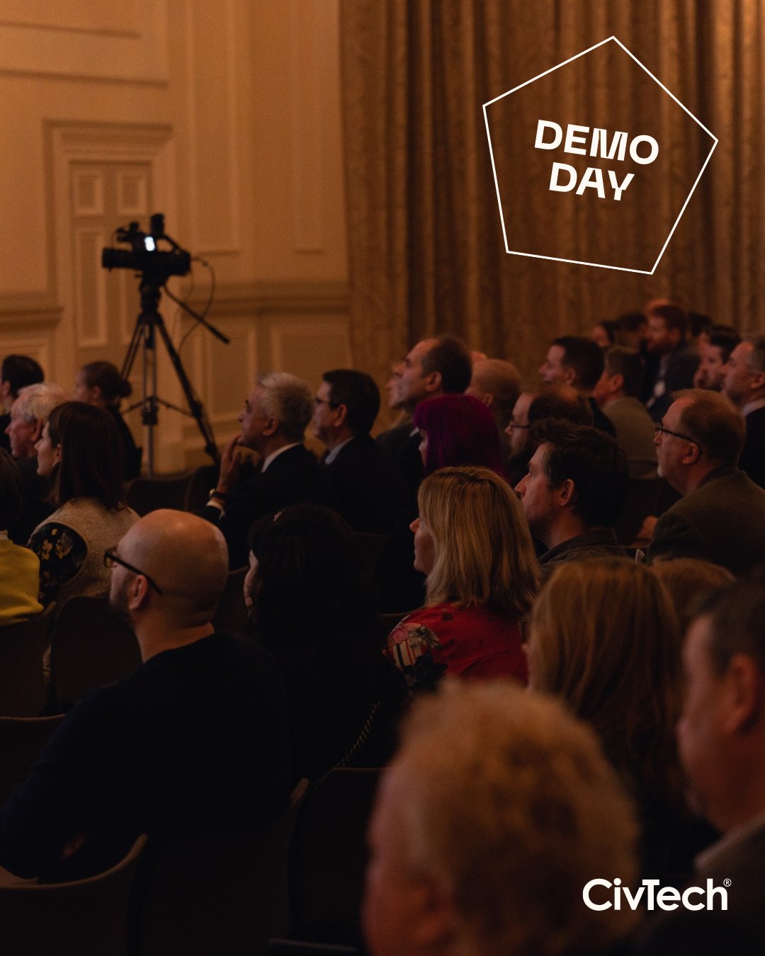 Dimly lit room with a crowd at Demo Day looking on towards the stage where companies are pitching. "Demo Day" text graphic overlay.