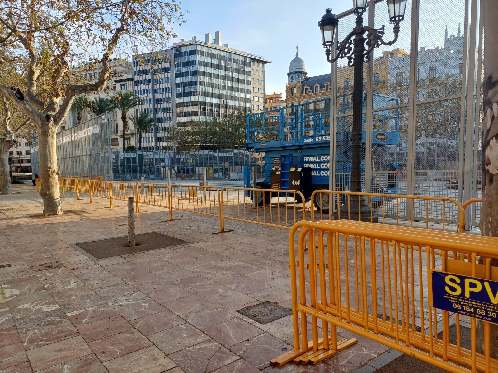 Barreras amarillas que cierran una zona peatonal junto a una plataforma elevadora azul, con árboles y edificios al fondo. Espacio central de la plaza de l'Ajuntament destinado al disparo de la mascletá en periodo fallero.