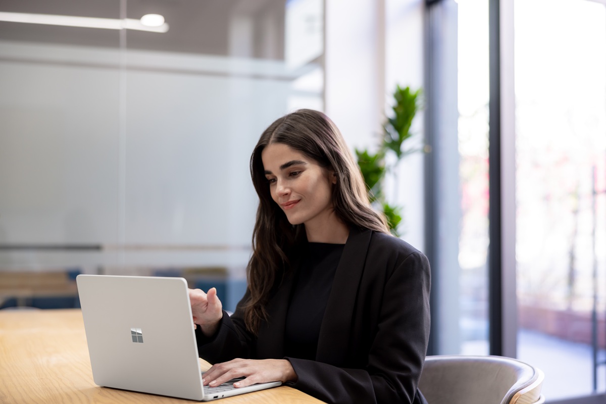 A person works on a Surface laptop at a table in a bright, modern office with glass walls and natural light.
