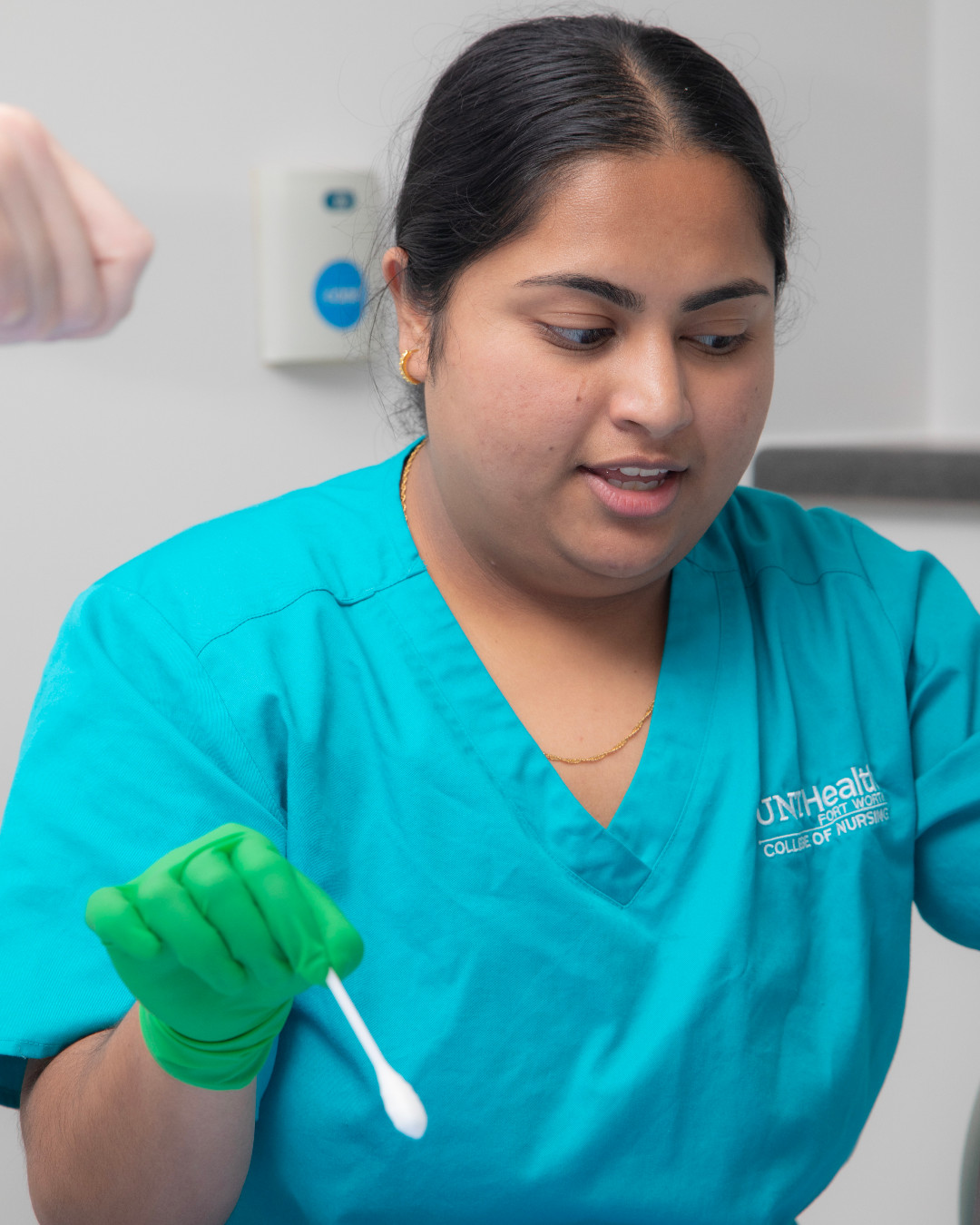 Sayeda Ali in teal medical scrubs and green gloves is holding a cotton swab in a clinical setting. The scrubs have "UNT Health College of Nursing" printed on them.