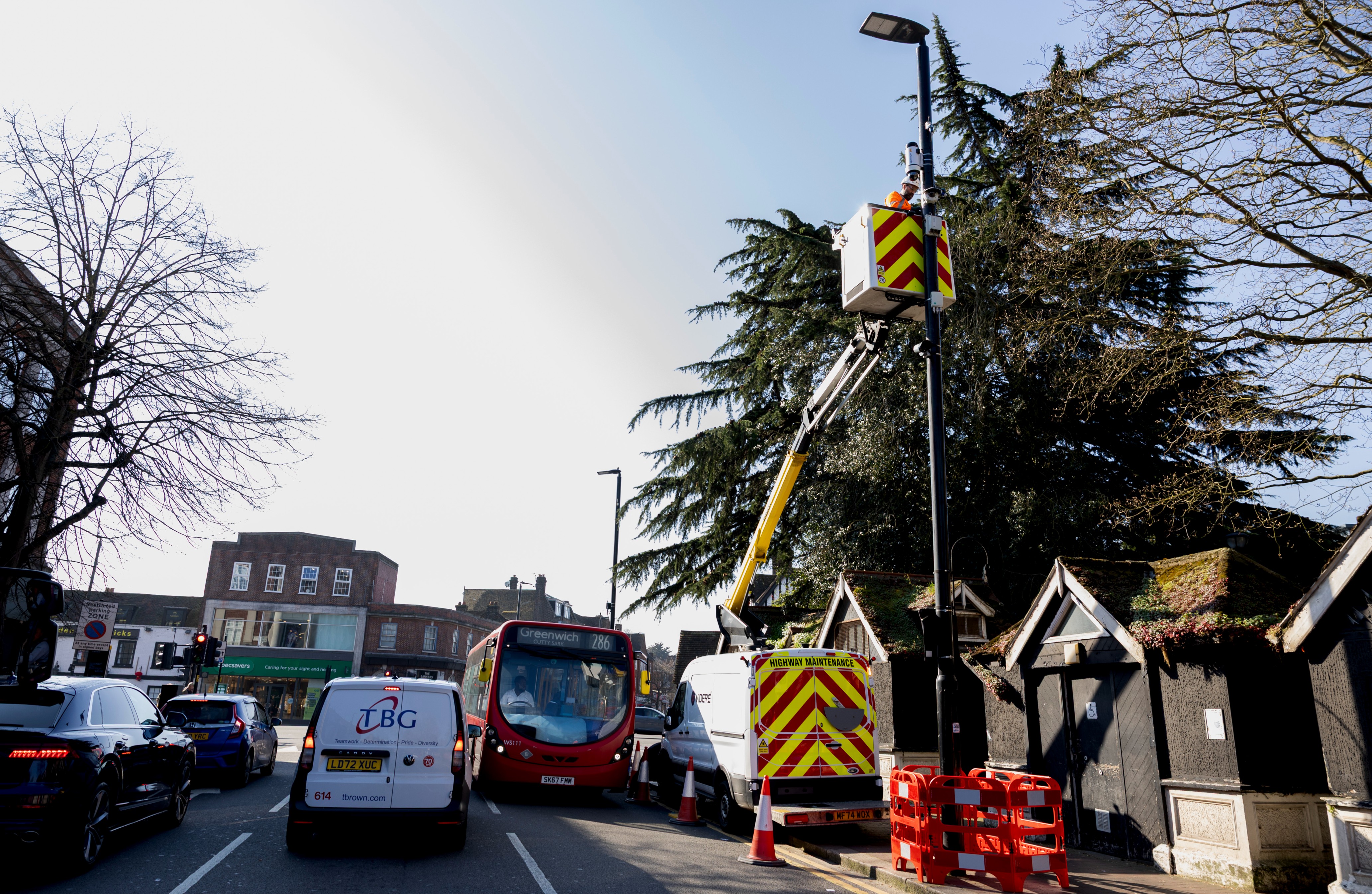 A worker inside a white, yellow-striped cherry picker attached to a highway maintenance truck is elevated on a lamppost in Eltham High Street installing a CCTV camera. 