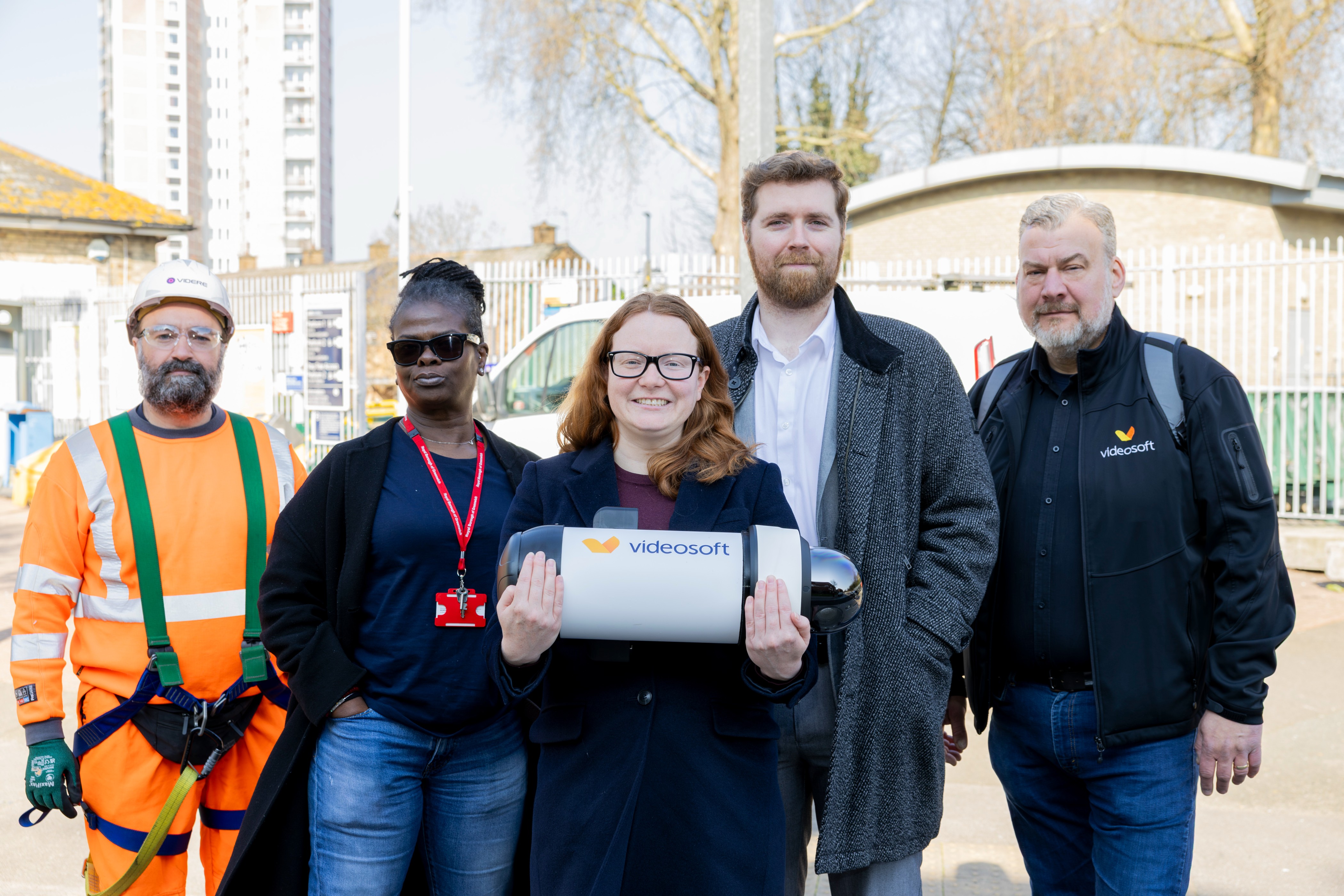 A group of five people stand outdoors in front of a fenced area. Cllr Taggart-Ryan holds a CCTV camera marked "videosoft." To the left is a person in an orange safety outfit and hard hat.