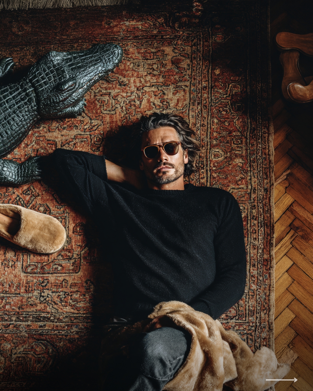Stylish man wearing sunglasses lying on a vintage rug beside a decorative crocodile sculpture in a cozy interior setting.