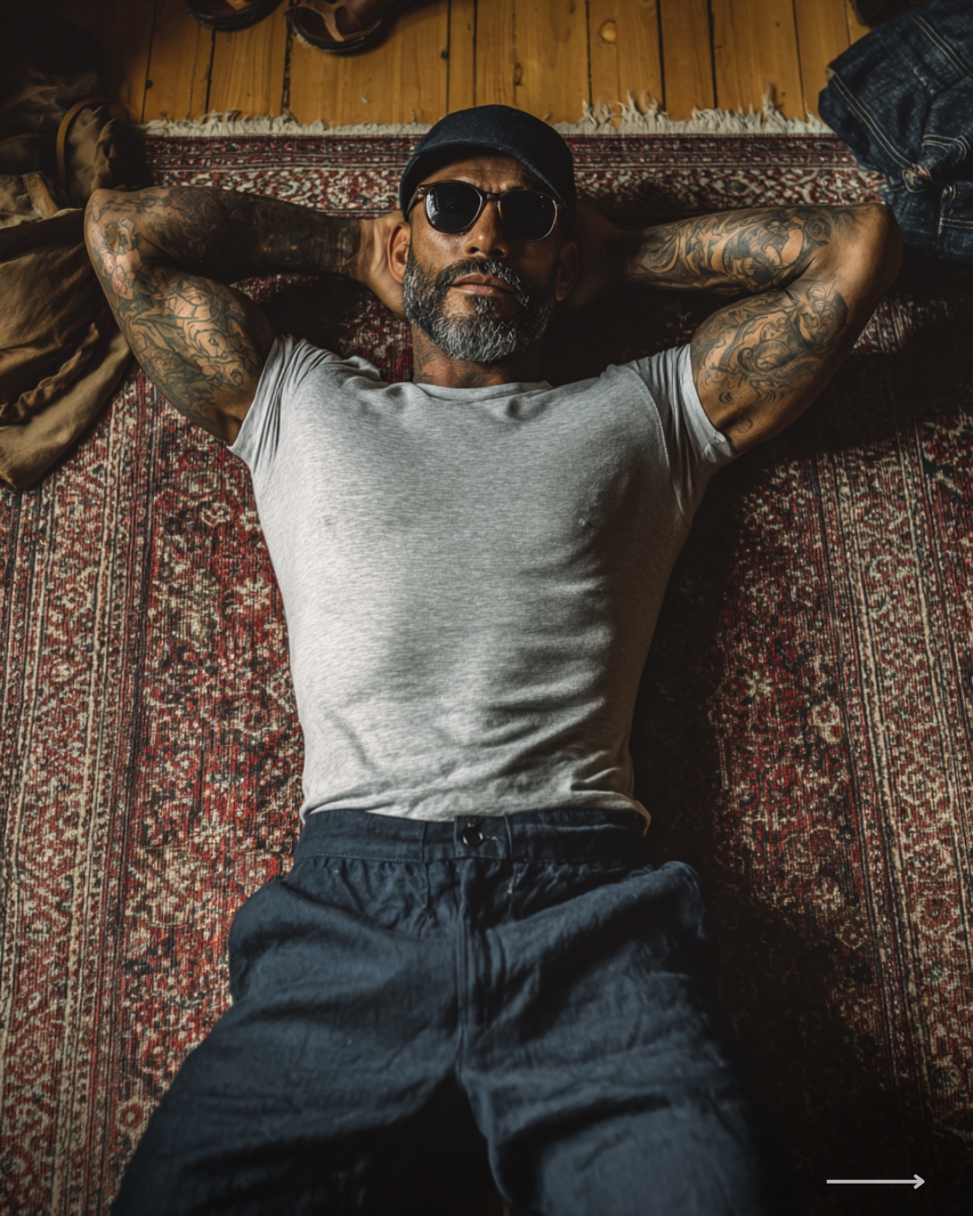 Tattooed man wearing sunglasses and a cap relaxing on a patterned rug with hands behind his head in a warm indoor setting.