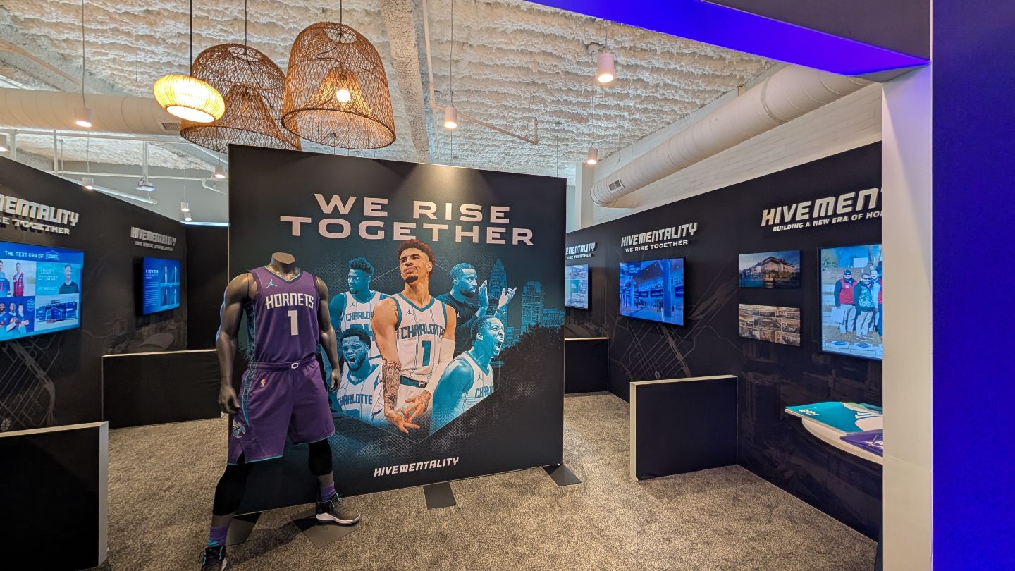 Hornets exhibit with a mannequin in a purple #1 jersey in front of a "We Rise Together" player mural and display screens