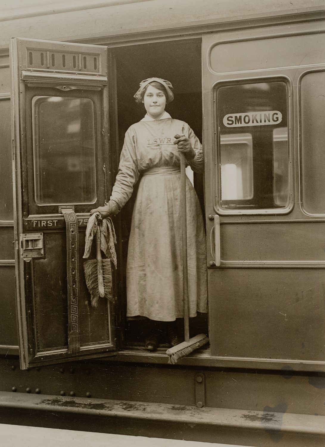 Woman in hat, protective gloves and overalls stood inside a railway carriage, holding a broom and cleaning materials, with the carriage door open. The overalls have 'LSWR' stitched in across the chest. Courtesy National Railway Museum.