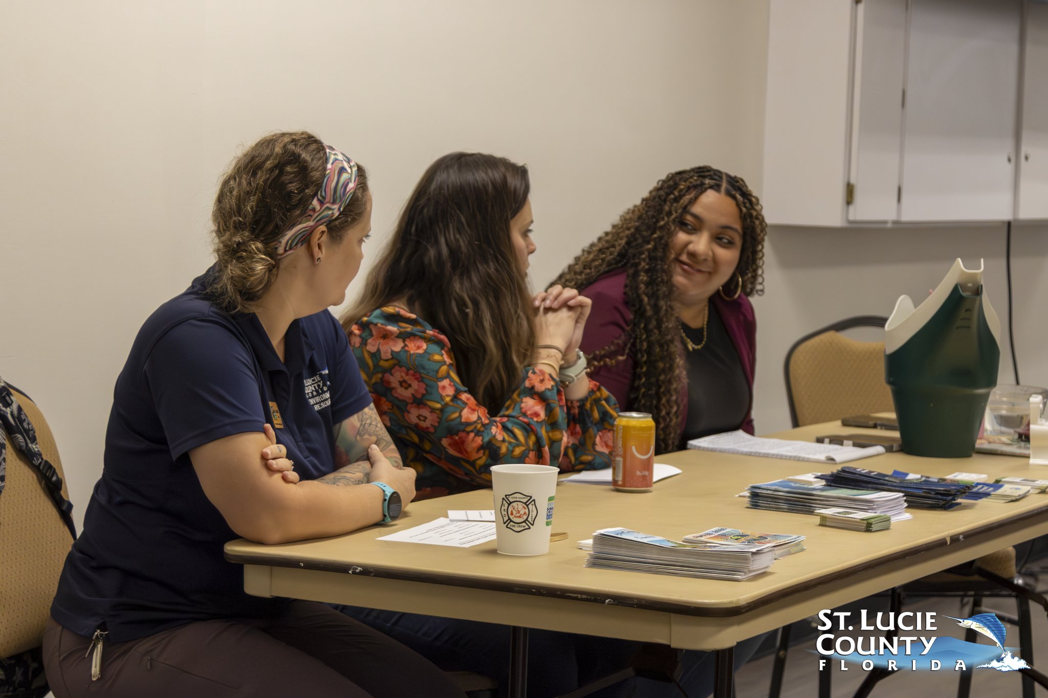 Three women seated at a table with informational brochures and drinks, engaged in discussion in a meeting room.