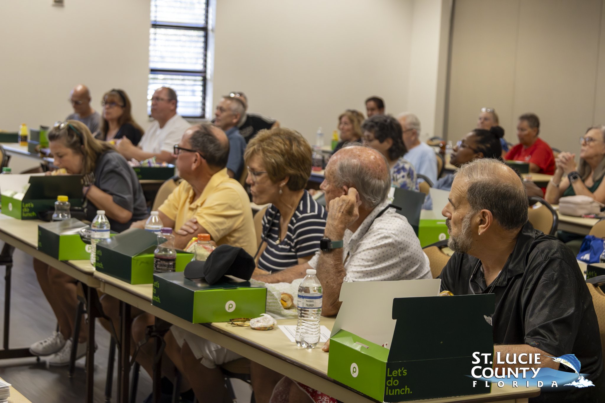 Adults attentively listening in a classroom setting with boxed lunches and water bottles on desks.