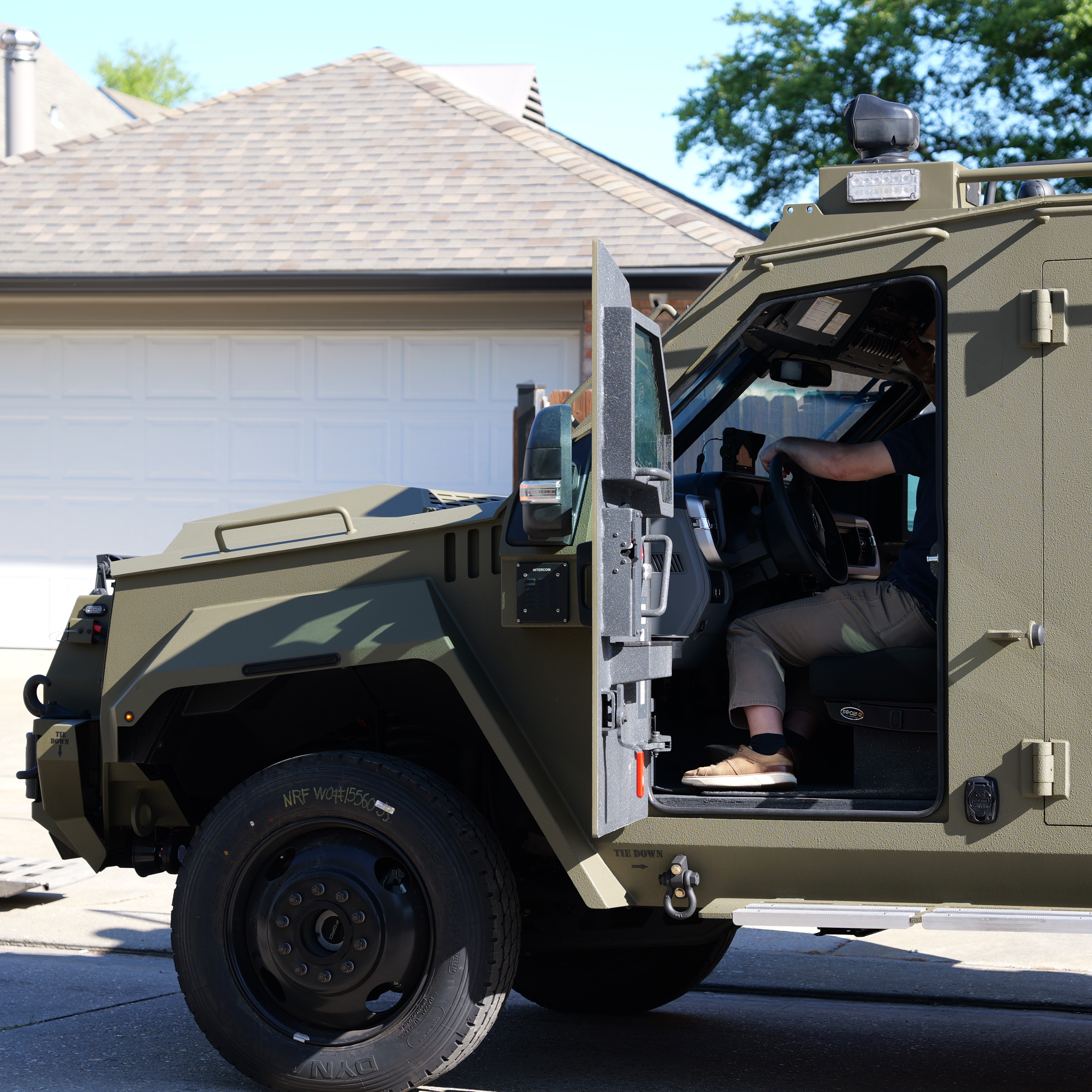 Person sitting inside an open-door military-style armored vehicle parked in a residential driveway.
