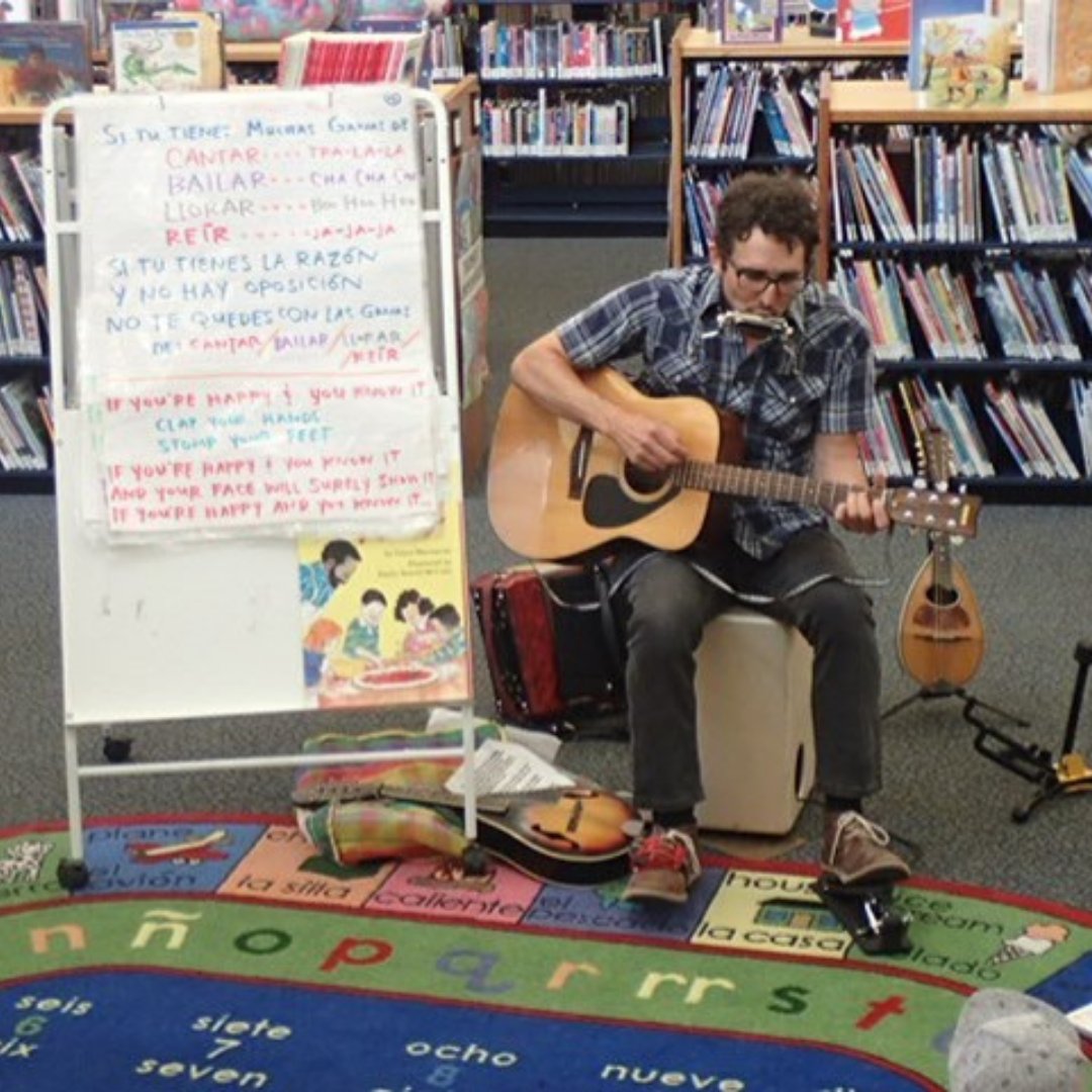 A person plays an acoustic guitar and a harmonica while sitting on a colorful rug in front of a whiteboard with writing and a bookshelf filled with books.