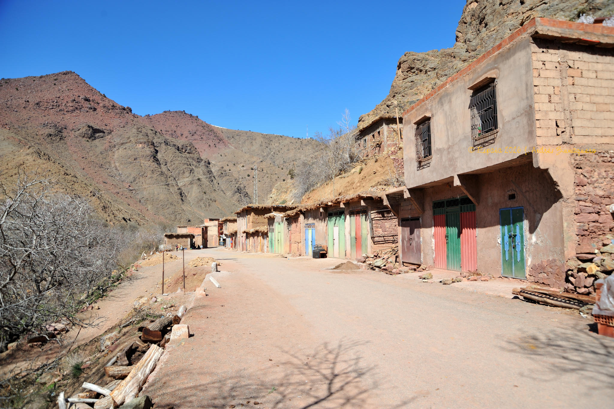 Bajo un cielo azul y fondo de una montaña, unas casas con sus puertas pintadas de colores vivos se alinean a lado derecho de la imagen frente a una calle cuyo pavimento es tierra del lugar.
