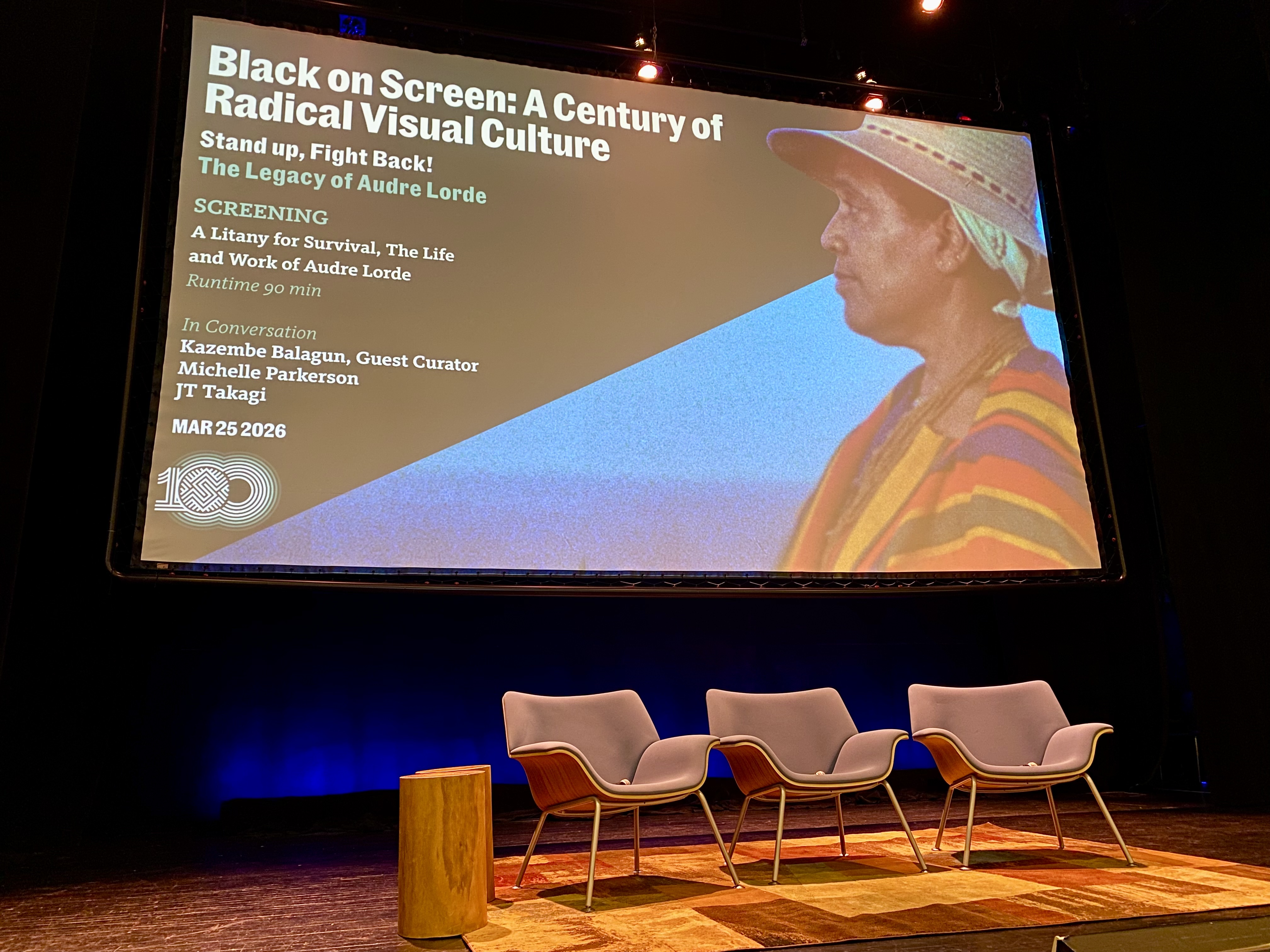 There is an empty stage with three modern chairs and a wooden stool beneath. Behind the chairs, there is a large screen displaying info the program's title "Black on Screen: A Century of Radical Visual Culture," date, and names of panelists.