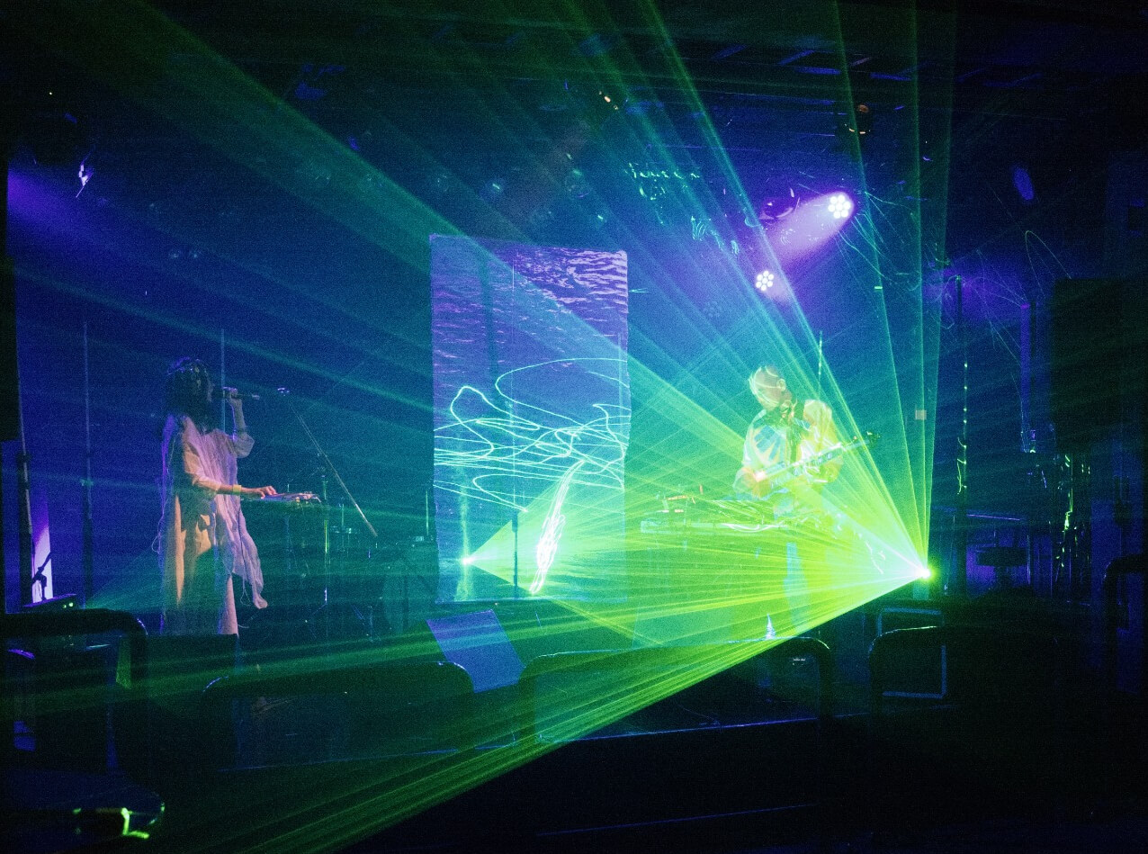 A live photo of Future Sound Lovers performing on a stage with lasers and LED lights. Kyra, the vocalist, is standing on the left and dressed in white clothing. Akira is standing on the right performing on his instrument behind a table.