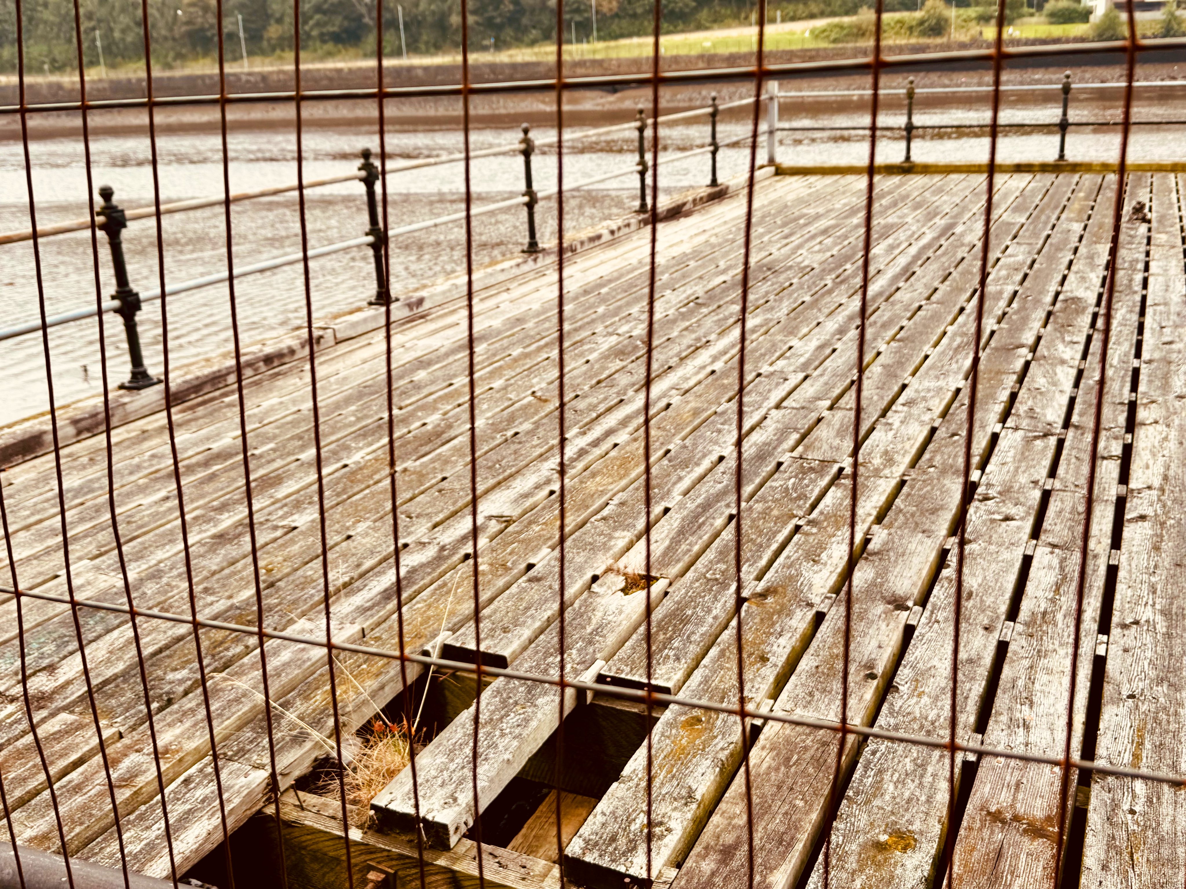 The remains of a wooden jetty, with parts of some of its planks missing. A metal fence put up to prevent members of the public from putting themselves in danger is now rusty.