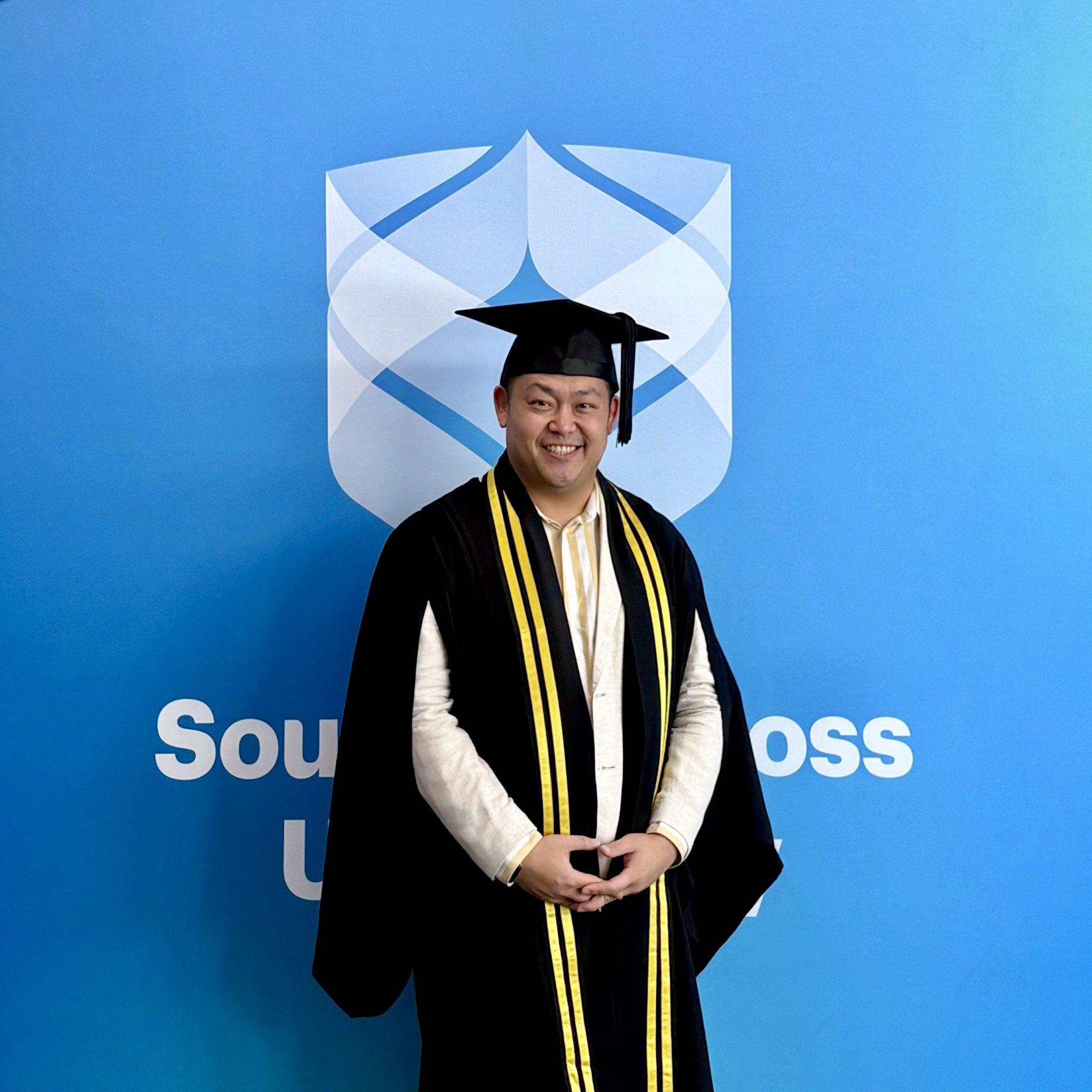 Leon Ha, standing in front of Southern Cross University photo wall, in his graduation regalia, celebrating the achievement of Graduate Certificate in Project Management.