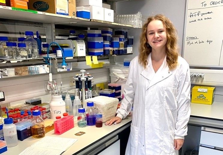 A scientist pictured wearing a white coat in a laboratory setting, with scientific equipment arranged on a workbench and shelving.