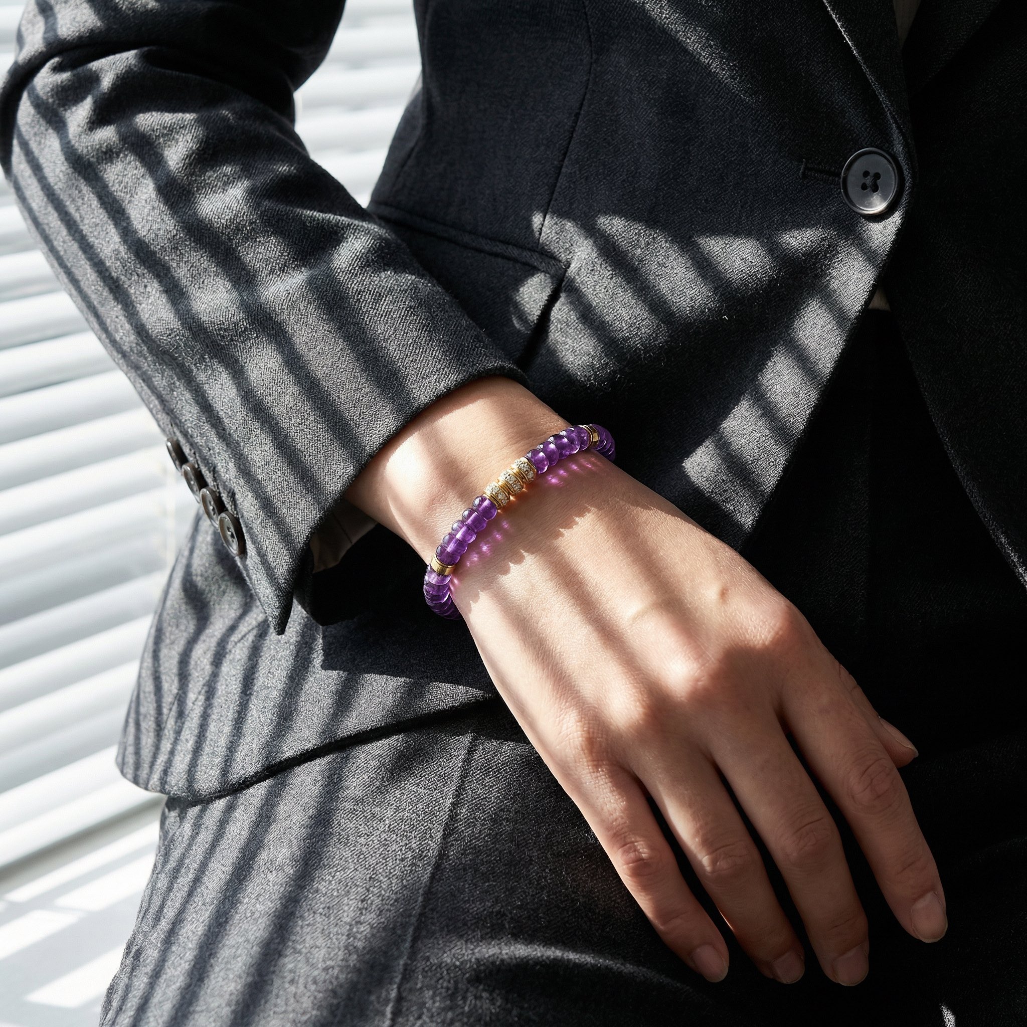 A close-up view focusing on a wrist wearing an exquisite amethyst beaded bracelet with multiple gold and diamond accents. The cuff of a textured charcoal grey suit jacket is visible. Dramatic, sharp striped shadows from window blinds cast a striking pattern across the skin.