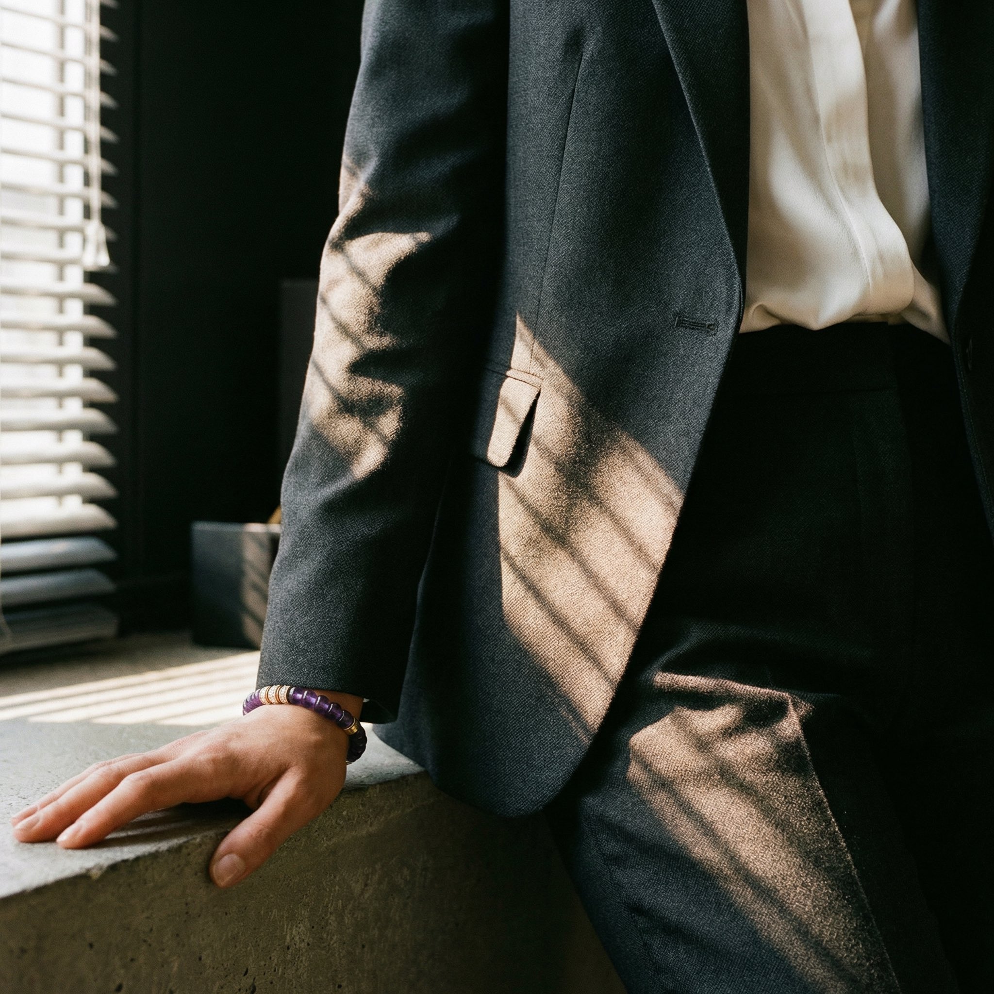 A person in a sharp, charcoal grey suit and crisp white shirt rests their hand on a concrete ledge. Their left wrist features a purple amethyst and single gold-diamond beaded bracelet. Dramatic, striped shadows from window blinds cover the entire scene.