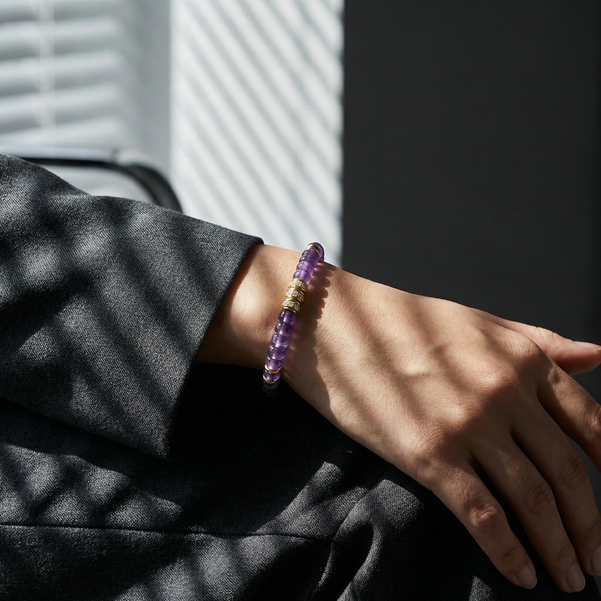 Close-up of a hand resting palm-down on textured grey suit fabric. The wrist is adorned with a purple amethyst and gold-diamond beaded bracelet. Strong, intricate, and highly graphic striped shadows from window blinds cast a complex zebra pattern across the entire hand and bracelet, making them the central focus.