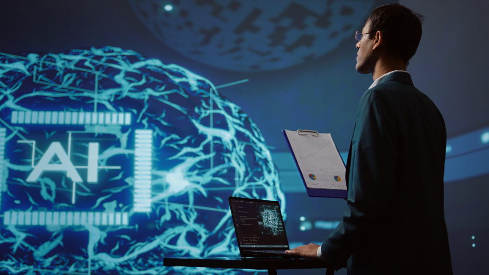 A man in a suit stands at a workstation with a laptop, holding a clipboard. Behind him is a large digital display featuring an abstract representation of a neural network and the text "AI." The setting appears to be a modern technological environment.