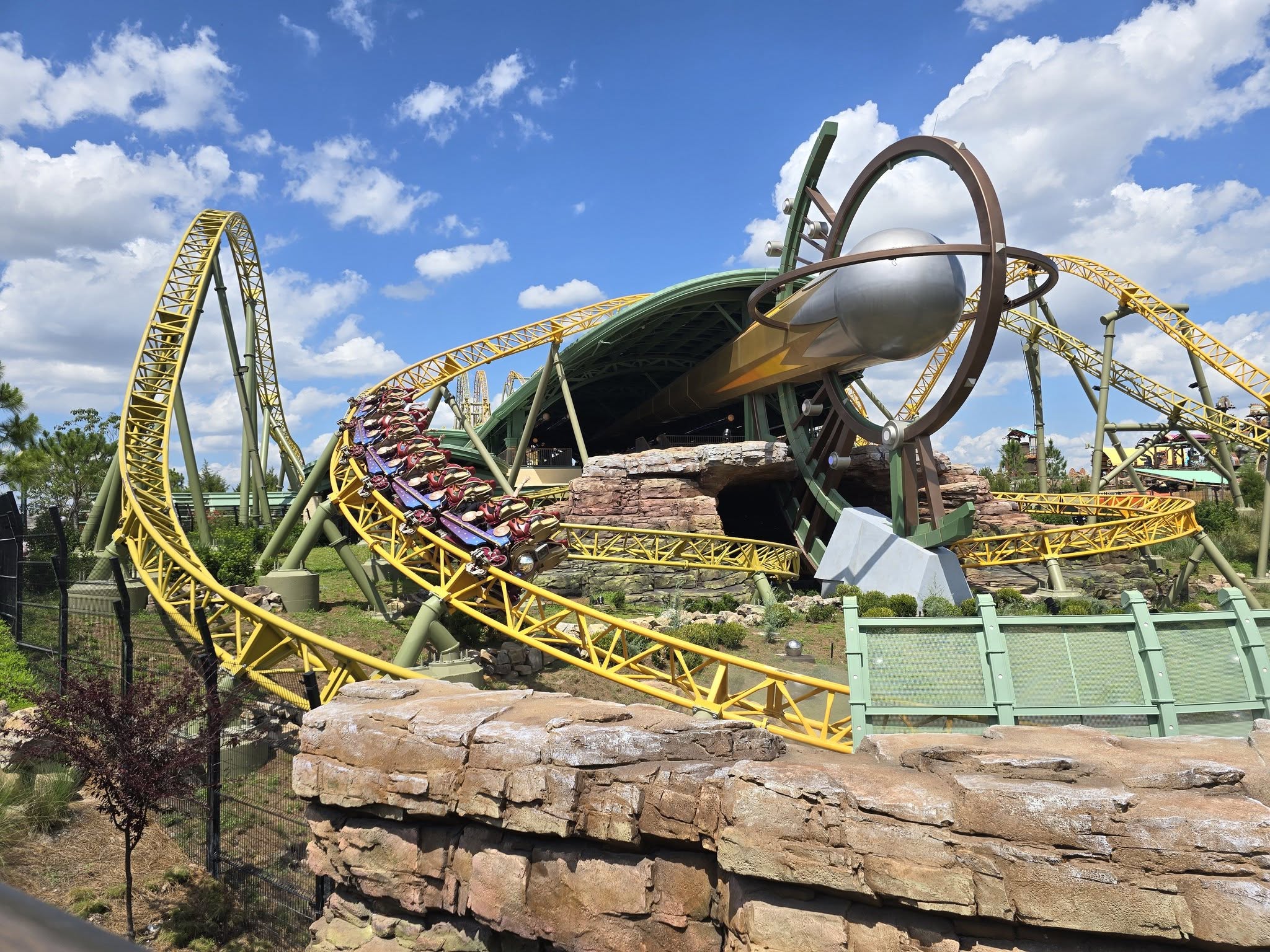A vibrant roller coaster at Universal Orlando's EPIC Universe is shown. The track is bright yellow with a complex loop, and a train full of people zooms along. The surrounding area features rocky terrain and green structures under a clear blue sky with scattered clouds.