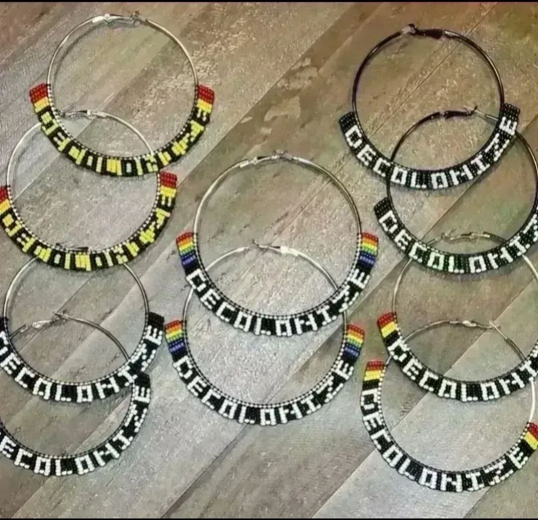 silver tone hoop earrings with various colors of beads, all say DECOLONIZE, displayed on a wood background 