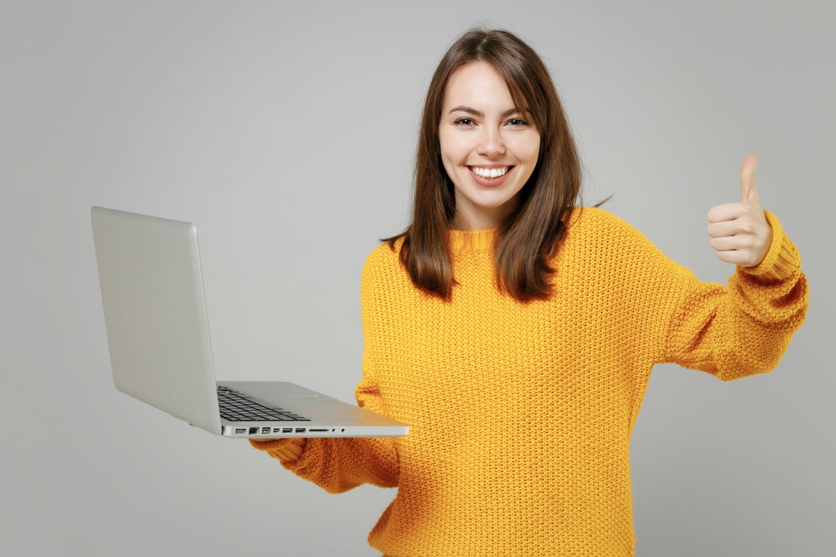 Young girl in a yellow sweater holding a laptop in one hand and giving a thumbs-up with the other.