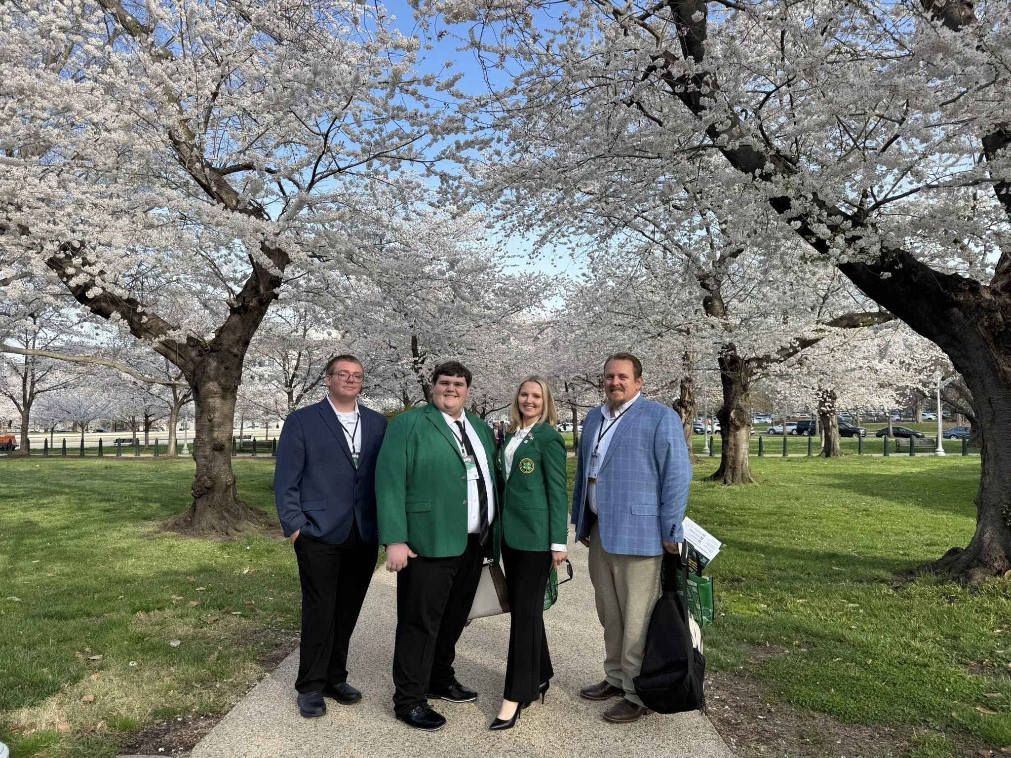 A group of four people stands on a path lined with cherry blossom trees. Two people wear green jackets. They are holding papers and credentials, suggesting a formal setting or event. The ground is grassy and well-maintained.