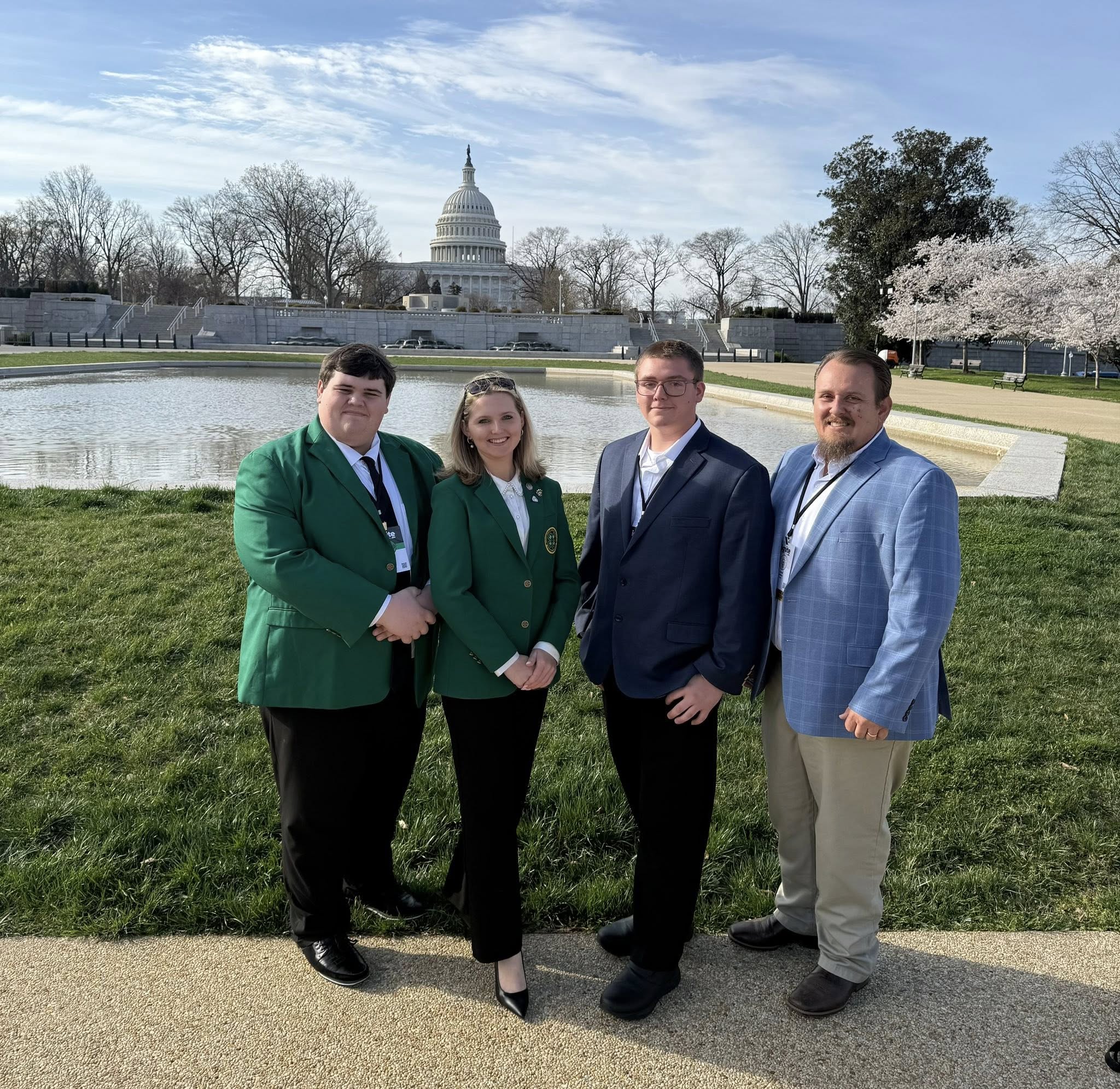 A group of four individuals stands outdoors in front of the United States Capitol building. They are dressed in formal attire, with two wearing green jackets. The scene includes a grassy area and a circular pond.