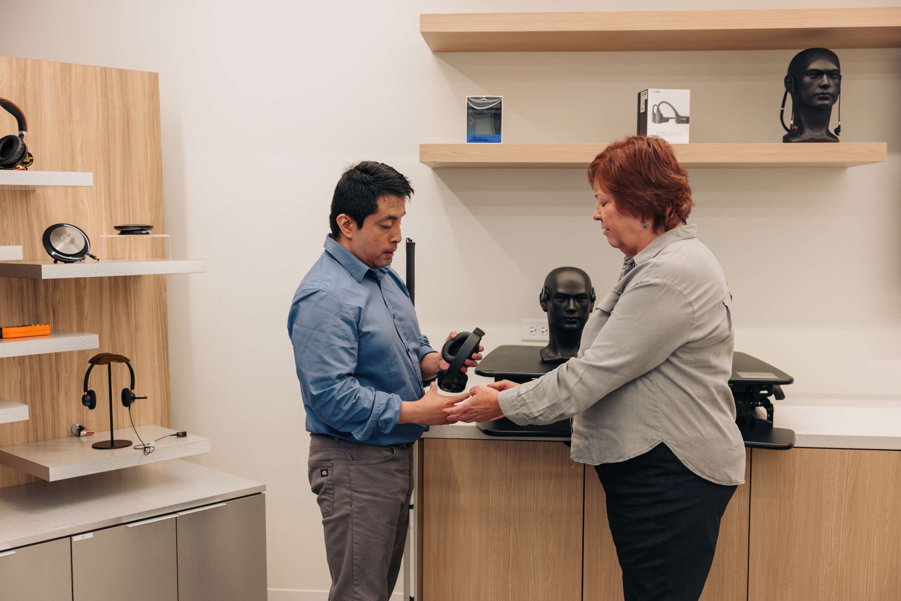 Deb and Israel in an Accessibility Solutions Center
Two professionals having a conversation in an accessibility solutions center. The woman holds out headphones to a man who is blind and holds his white cane against his body.

Photo Credit: Disability:IN