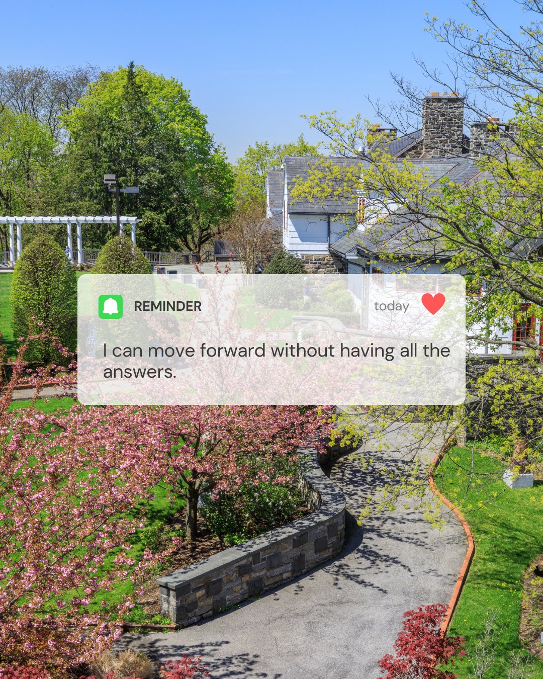 A winding garden path bordered by blooming pink and red trees leads past stone walls toward white houses under a clear blue sky with a reminder bubble that says, " I can move forward without having all the answers."