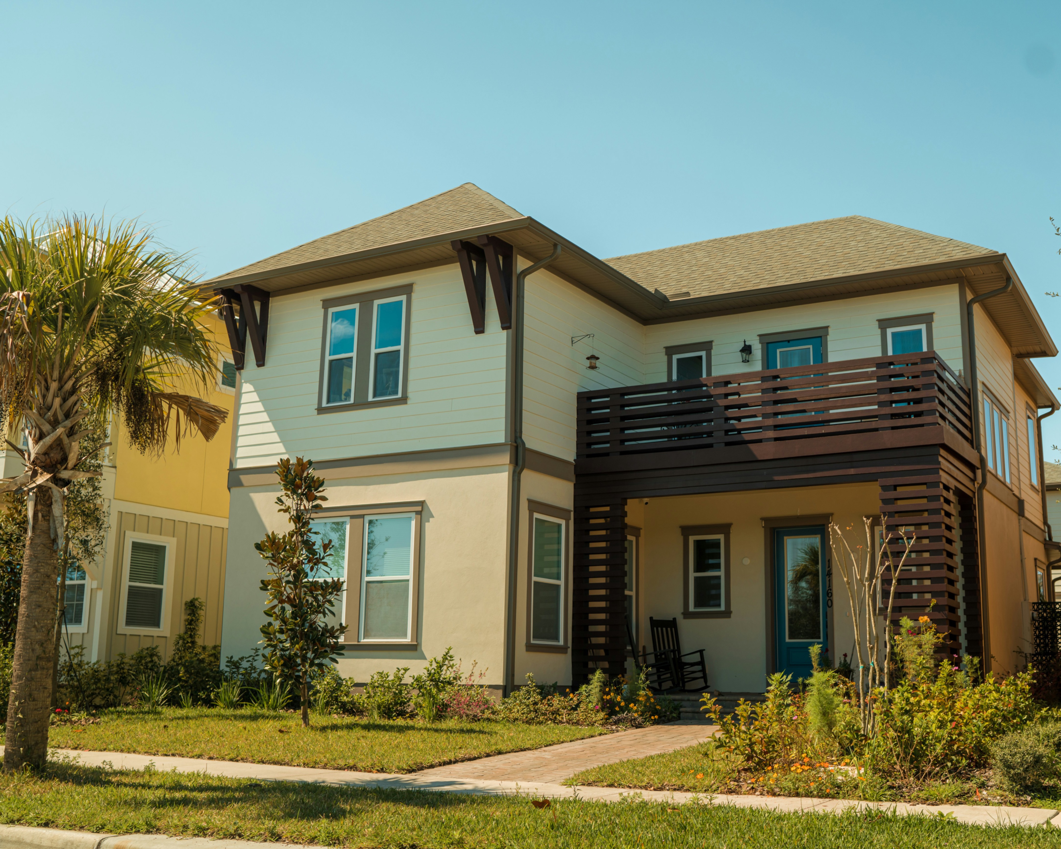 Modern two-story suburban home with a balcony, landscaped yard, and palm tree in a sunny neighborhood, representing comfortable and affordable housing.