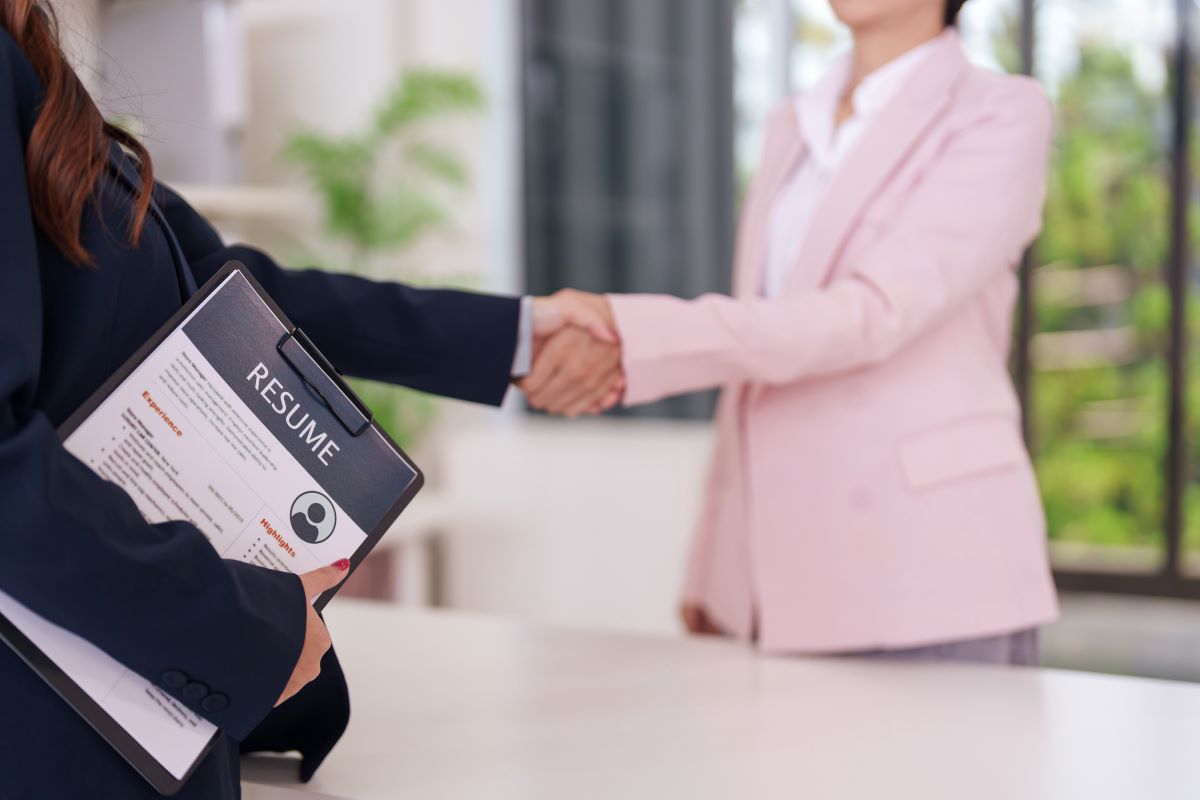 Two people shaking hands across a desk during a job interview, one holding a resume folder.