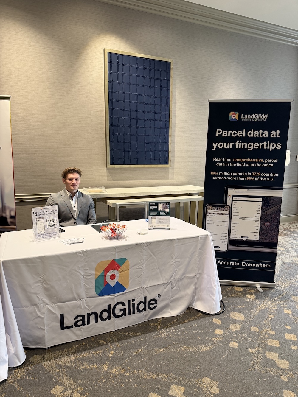 A representative sits at a booth displaying LandGlide materials. The table has a LandGlide-branded cloth, informational brochures, and a bowl of candy. A banner beside the booth highlights the features of LandGlide, emphasizing parcel data access.