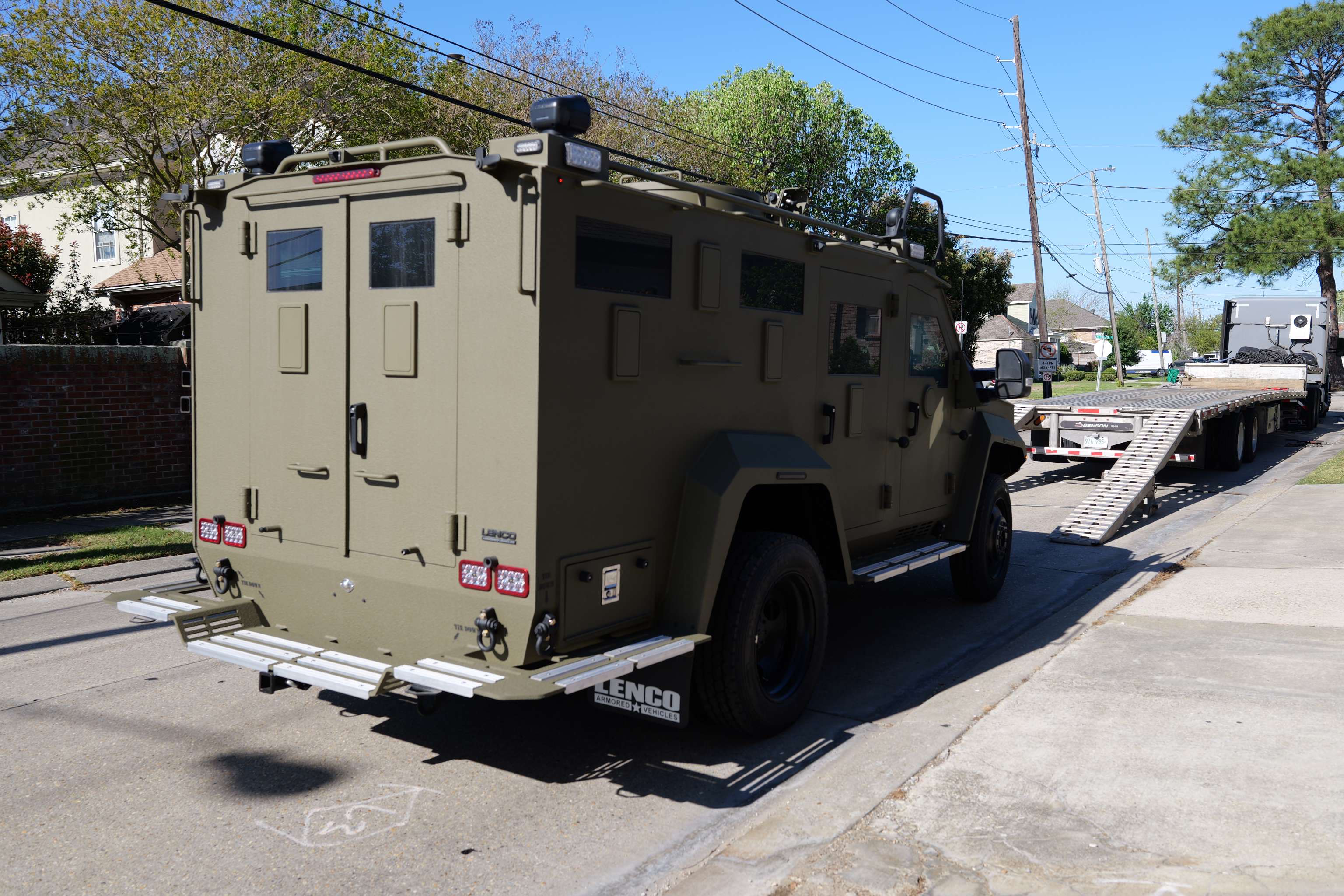 Tan armored vehicle parked on a residential street near a flatbed trailer with loading ramps.