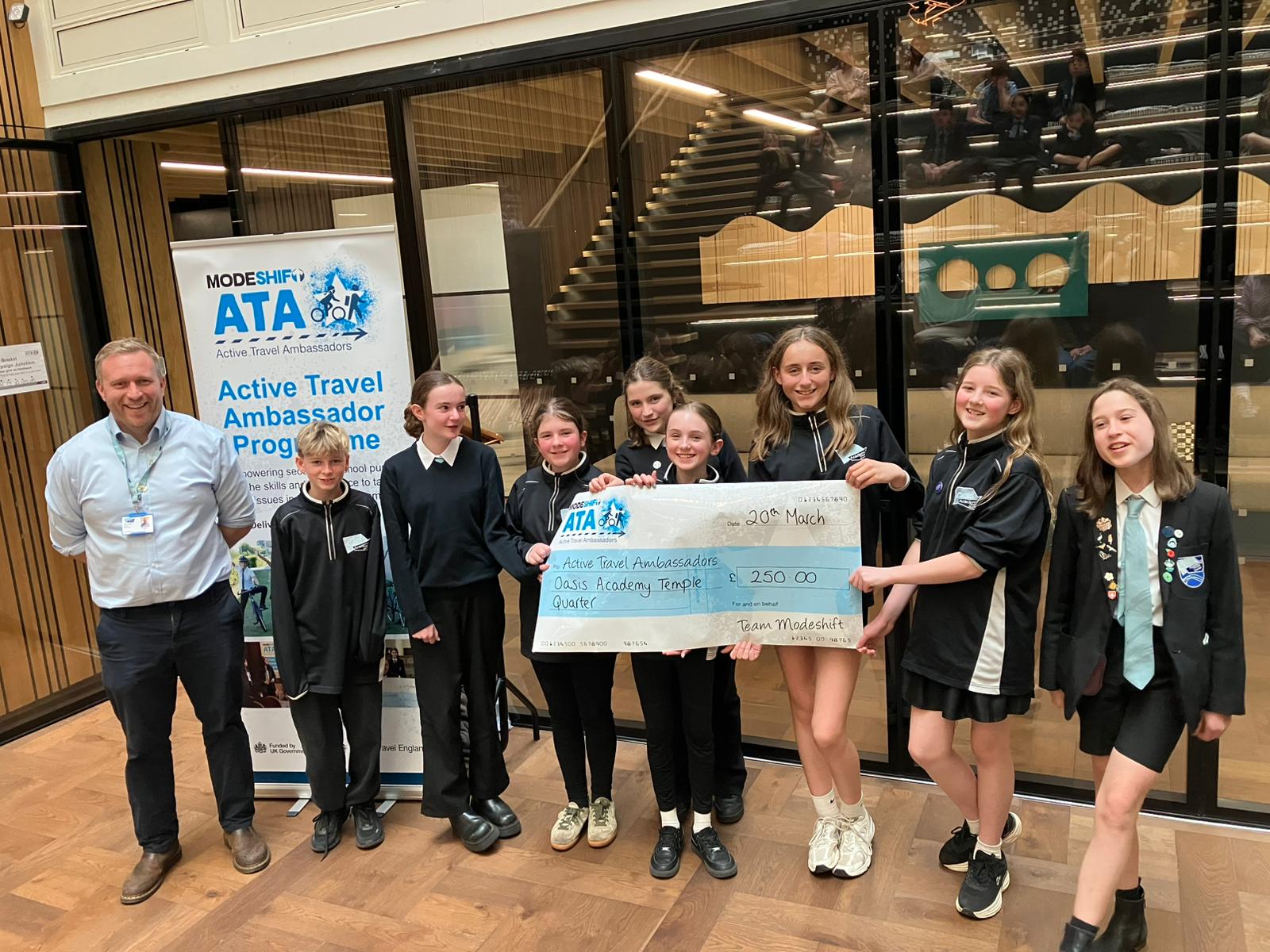Group of young students and an adult holding a large check for £250 from the Active Travel Ambassador Programme indoors.